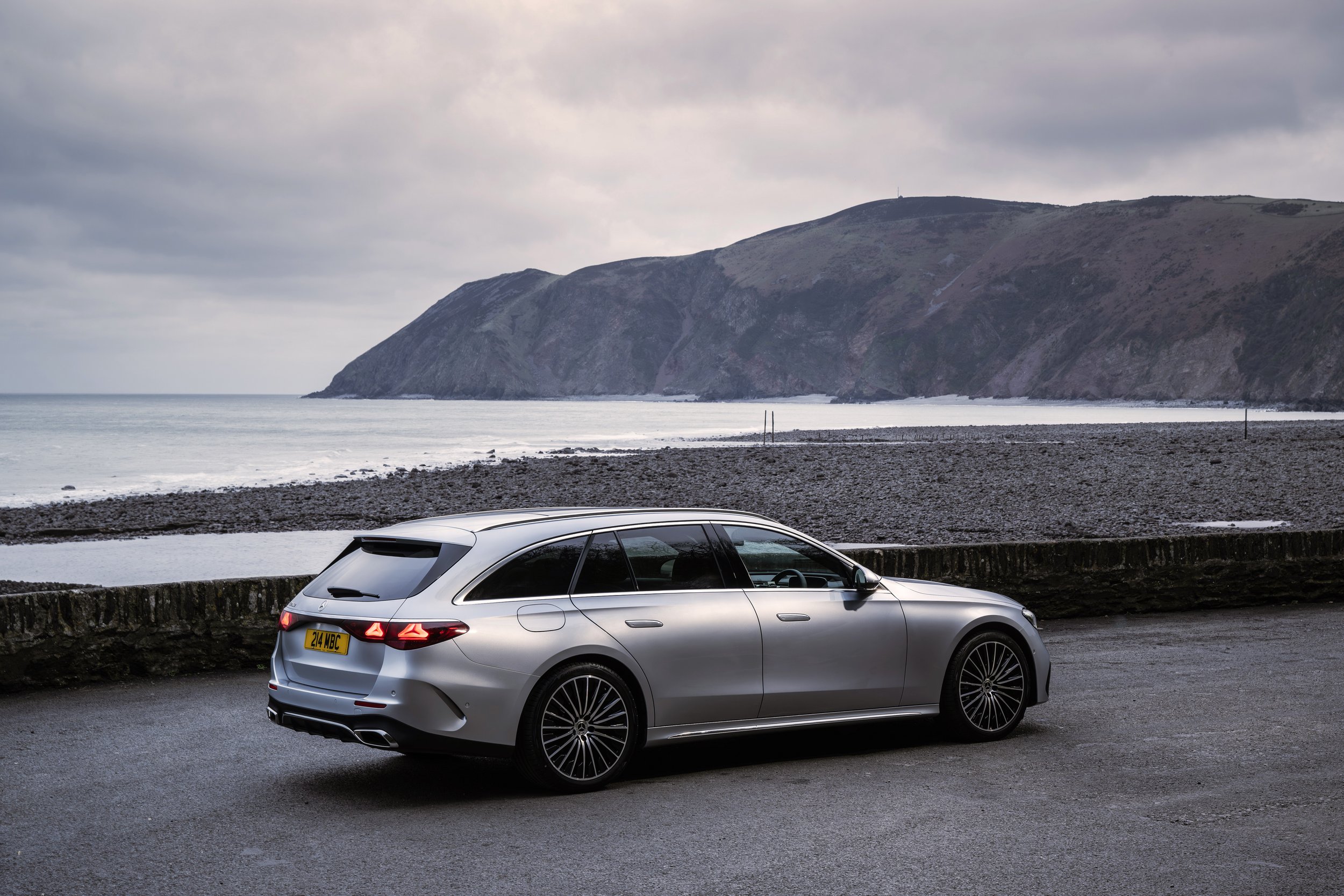 Silver luxury station wagon parked near a coastal shoreline with cliffs and ocean in the background, overcast sky.