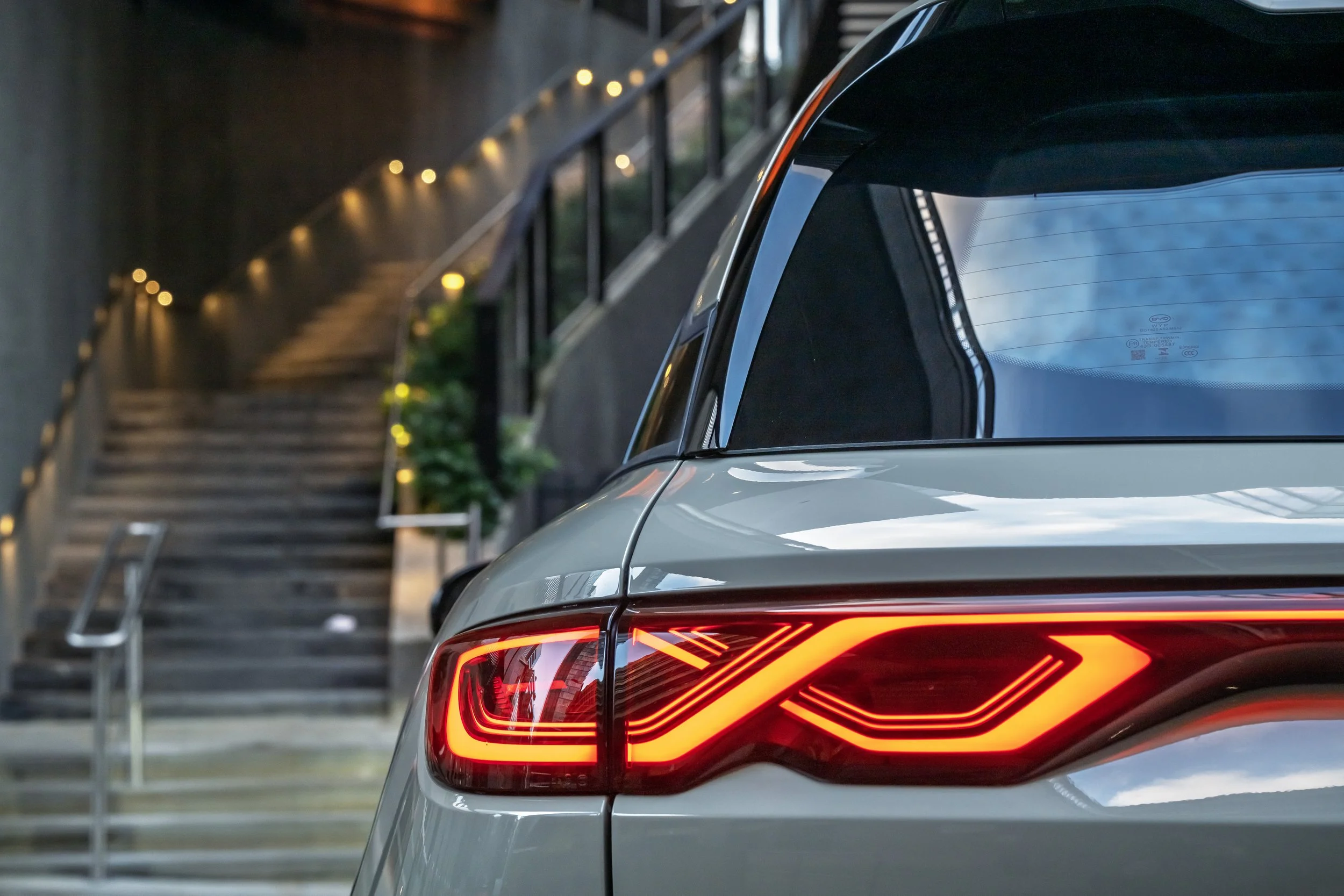 Close-up of rear end of a modern white SUV, showing its illuminated taillight. In the background, a staircase with warm lighting and railings, along with some greenery and indoor plants, is visible.