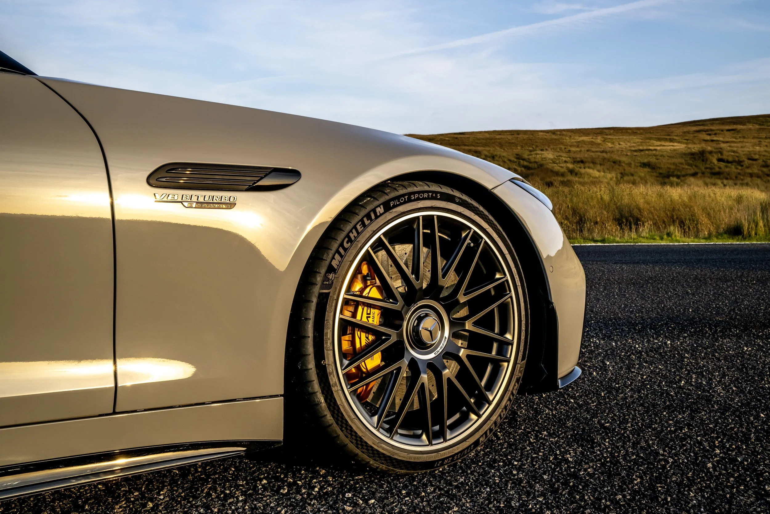 Close-up of the front part of a silver Mercedes-Benz car, showing a V8 biturbo badge, a large black alloy wheel with Michelin Pilot Sport 4S tire, and a yellow brake caliper, on a road with a grassy landscape in the background during sunset.