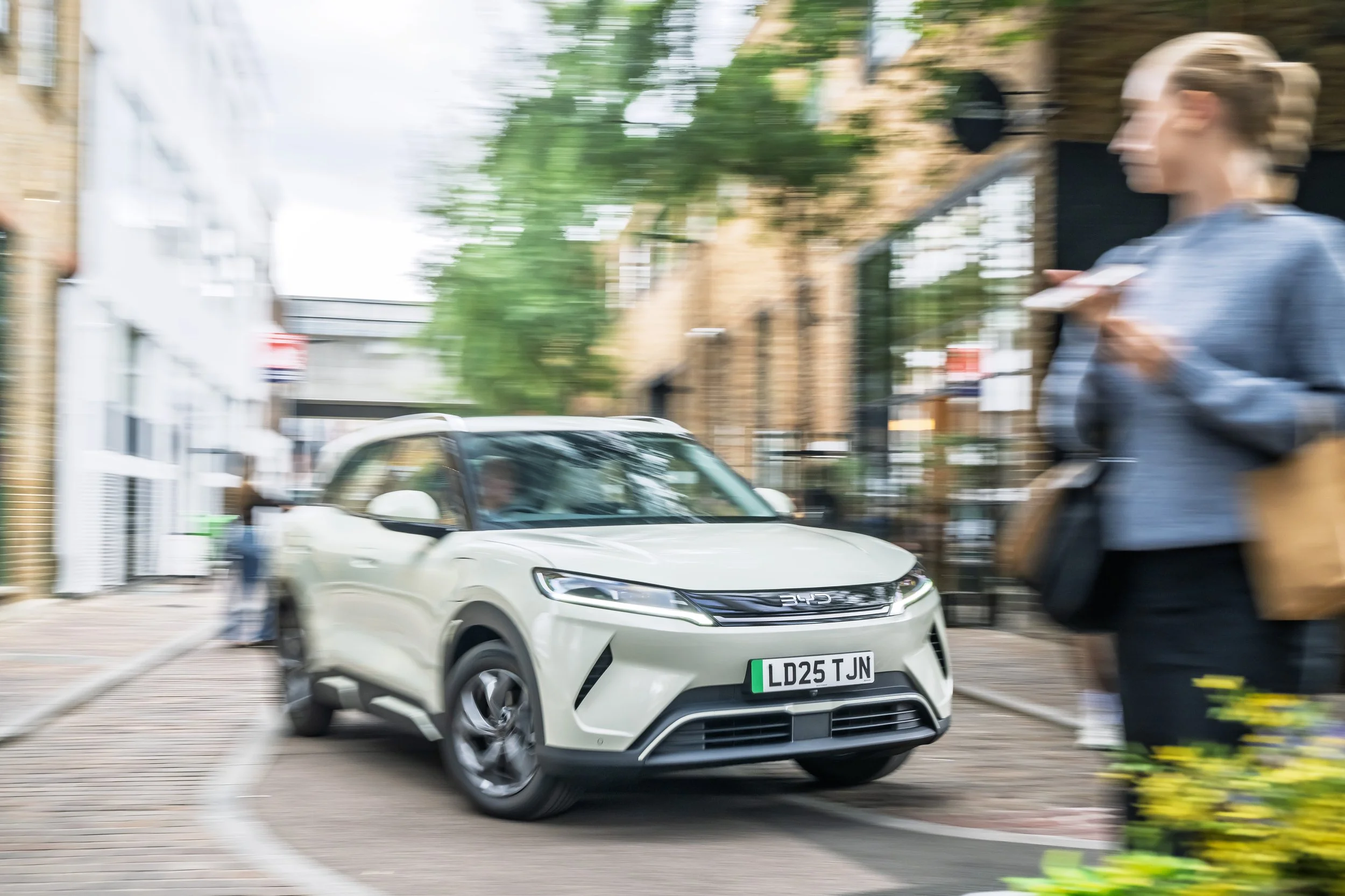 A white car driving on a city street, with a woman walking on the sidewalk next to it, holding a coffee cup.
