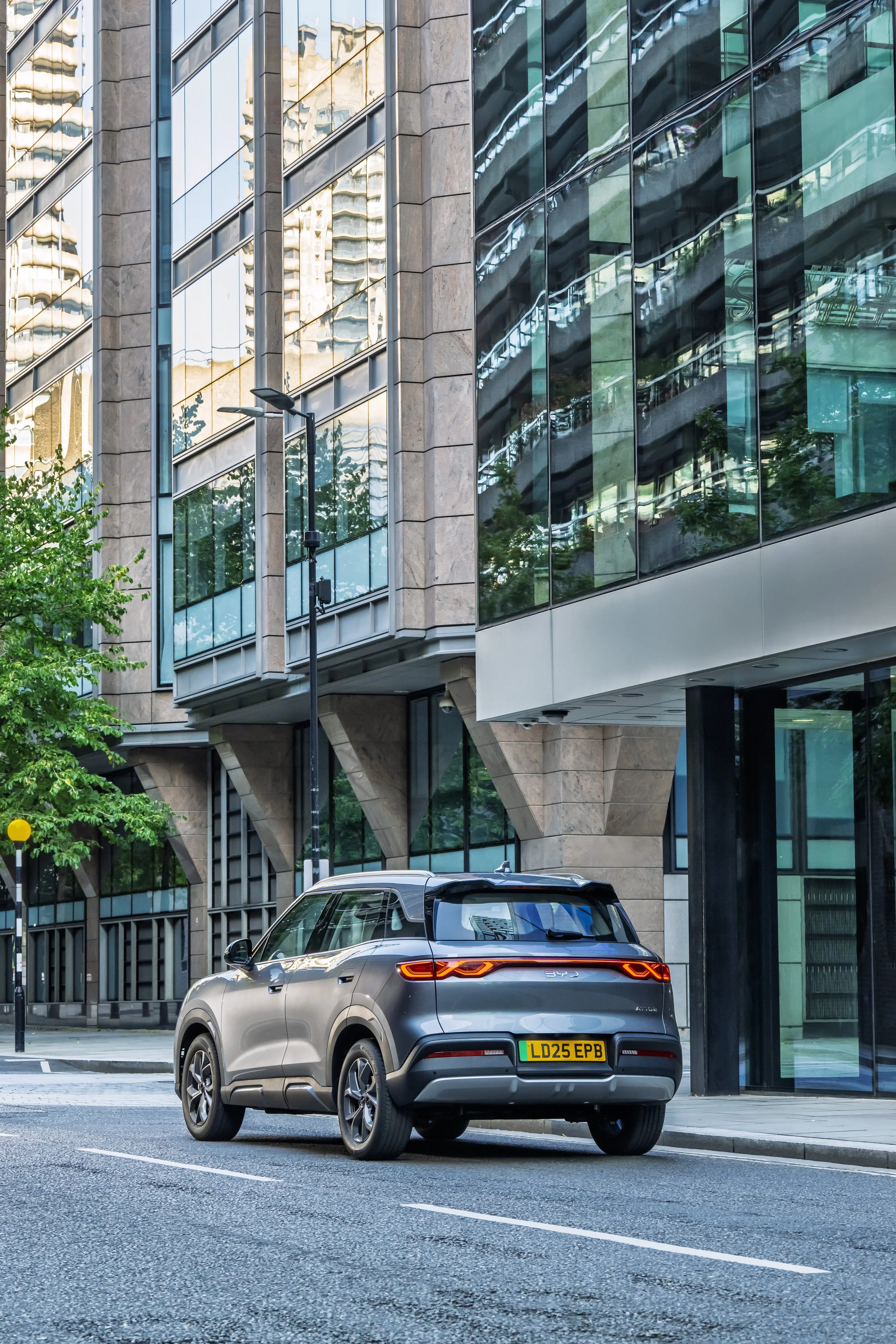 A gray SUV parked on a city street in front of modern glass and stone office buildings, with a green tree on the left side and a street lamp in the background.