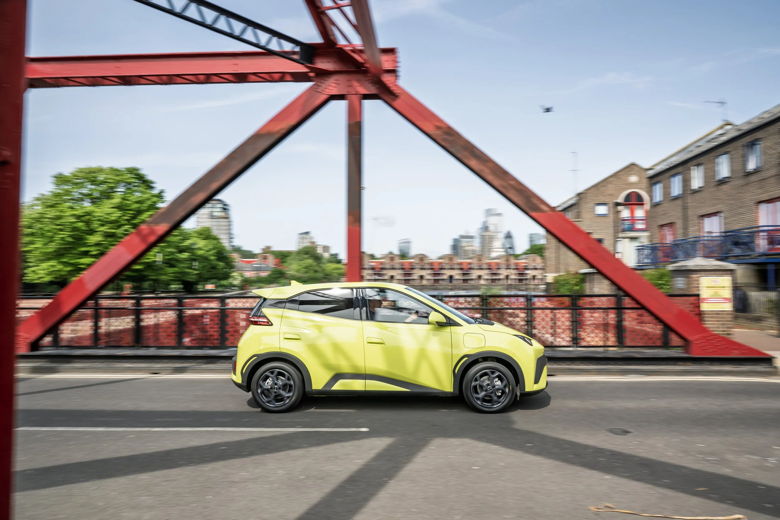 A small yellow electric car driving across a red bridge on a city street during daytime.