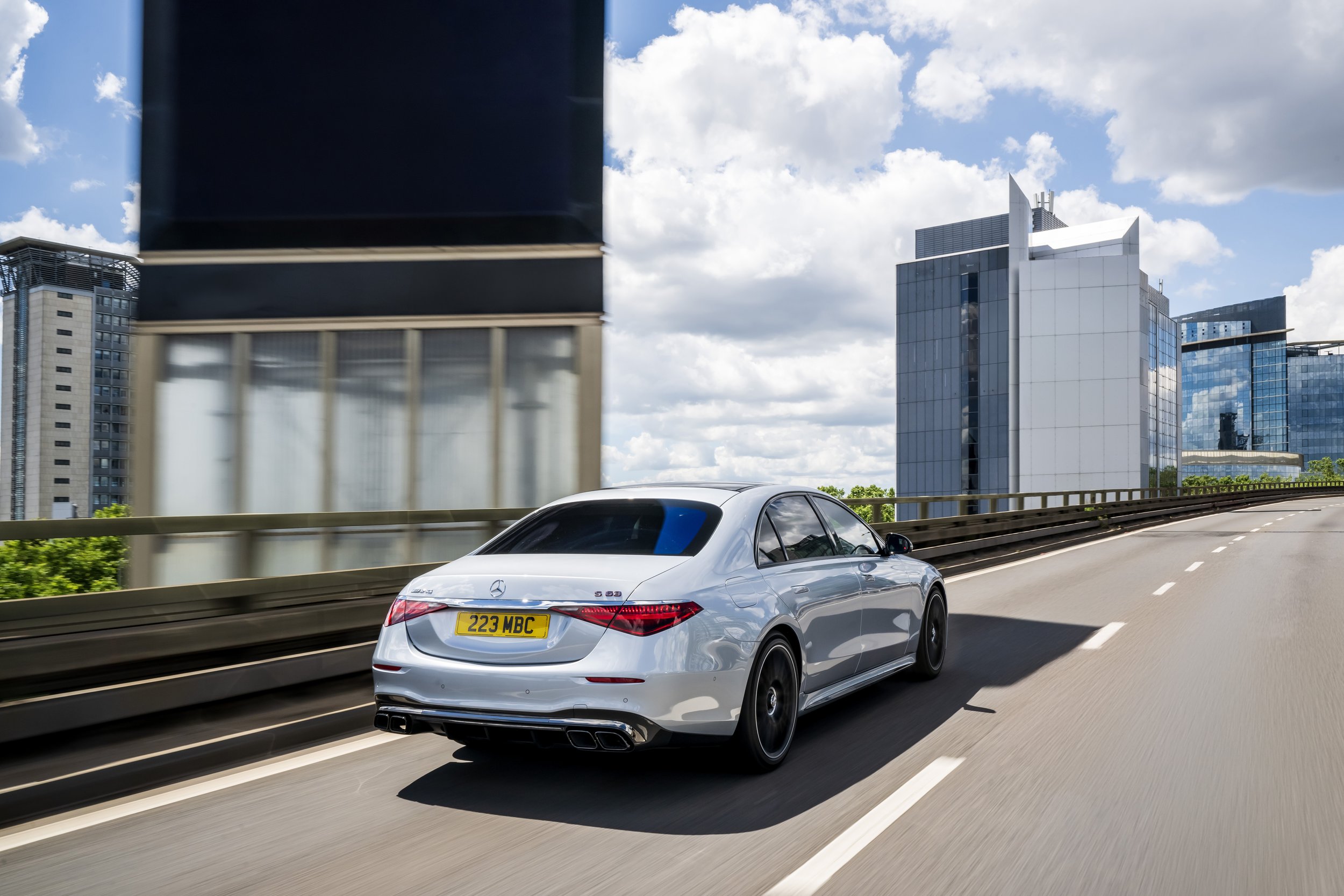 A silver Mercedes-Benz S-Class car driving on a highway with modern office buildings in the background on a partly cloudy day.
