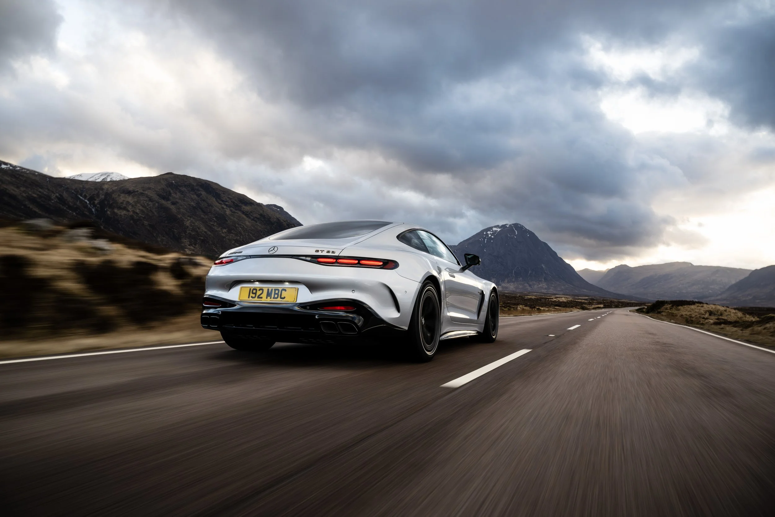 Silver Mercedes-Benz AMG GT sports car driving on open highway through mountainous landscape under cloudy sky.