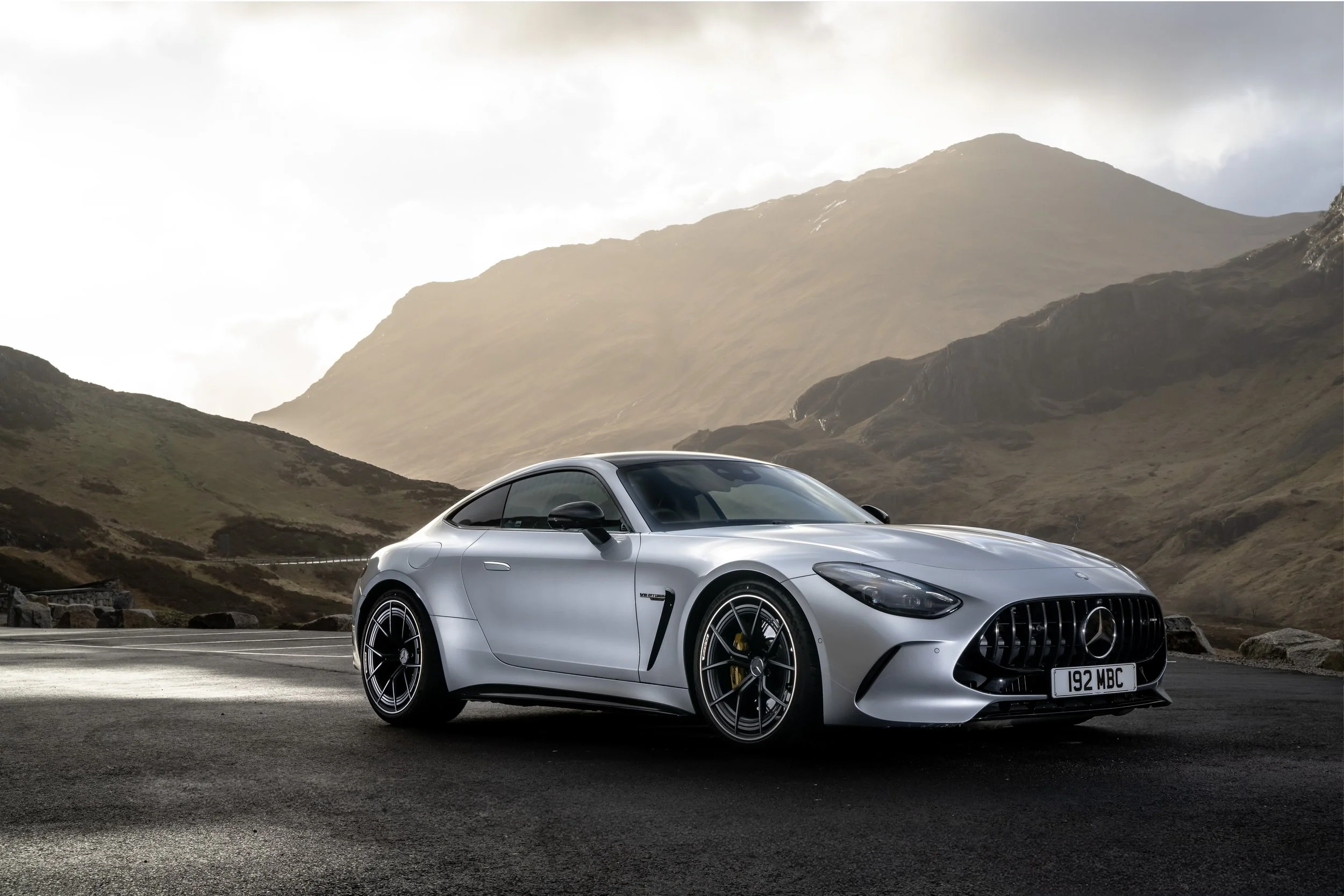 A silver Mercedes-Benz AMG GT sports car parked on a mountain road with a backdrop of hills and mountains under a cloudy sky.