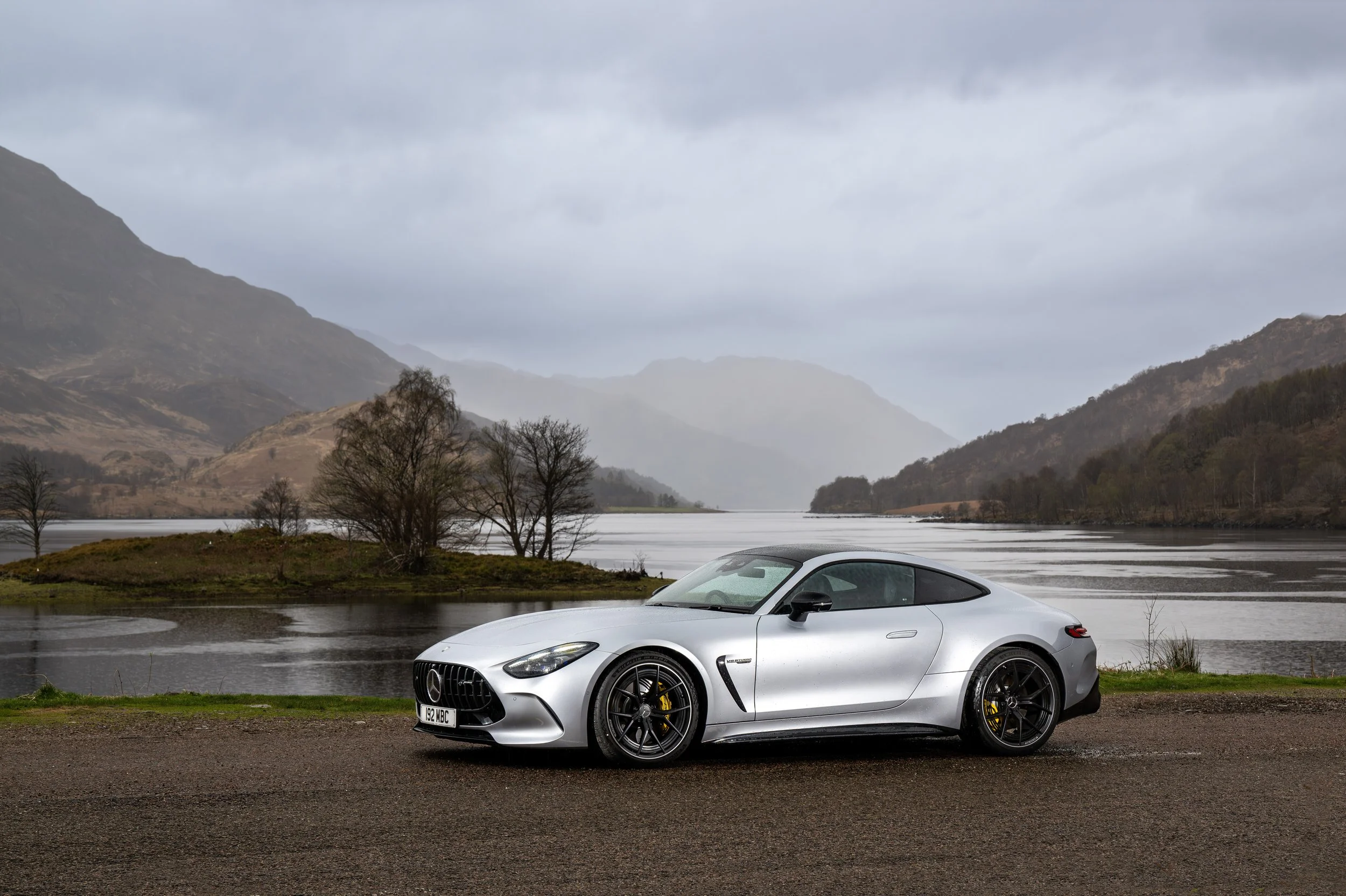 A silver sports car parked on a road by a lake with mountains in the background under a cloudy sky.