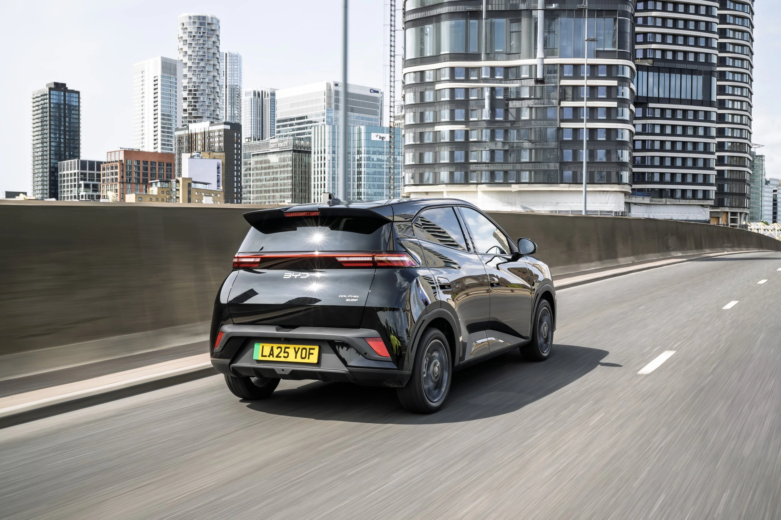 Black compact electric car driving on a city highway with tall modern buildings in the background.