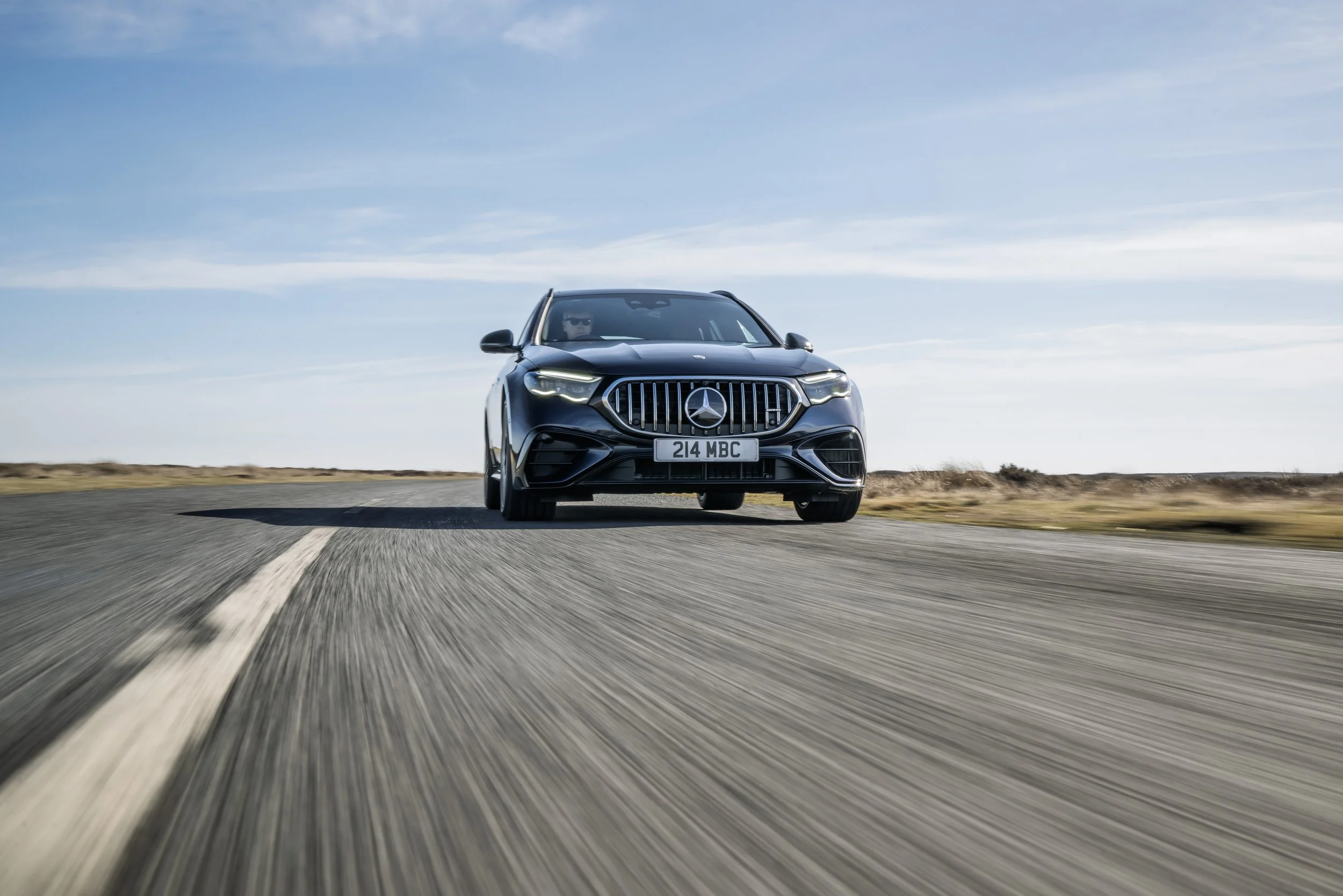 A black Mercedes-Benz car driving on a curved road in an open landscape under a sky with scattered clouds.