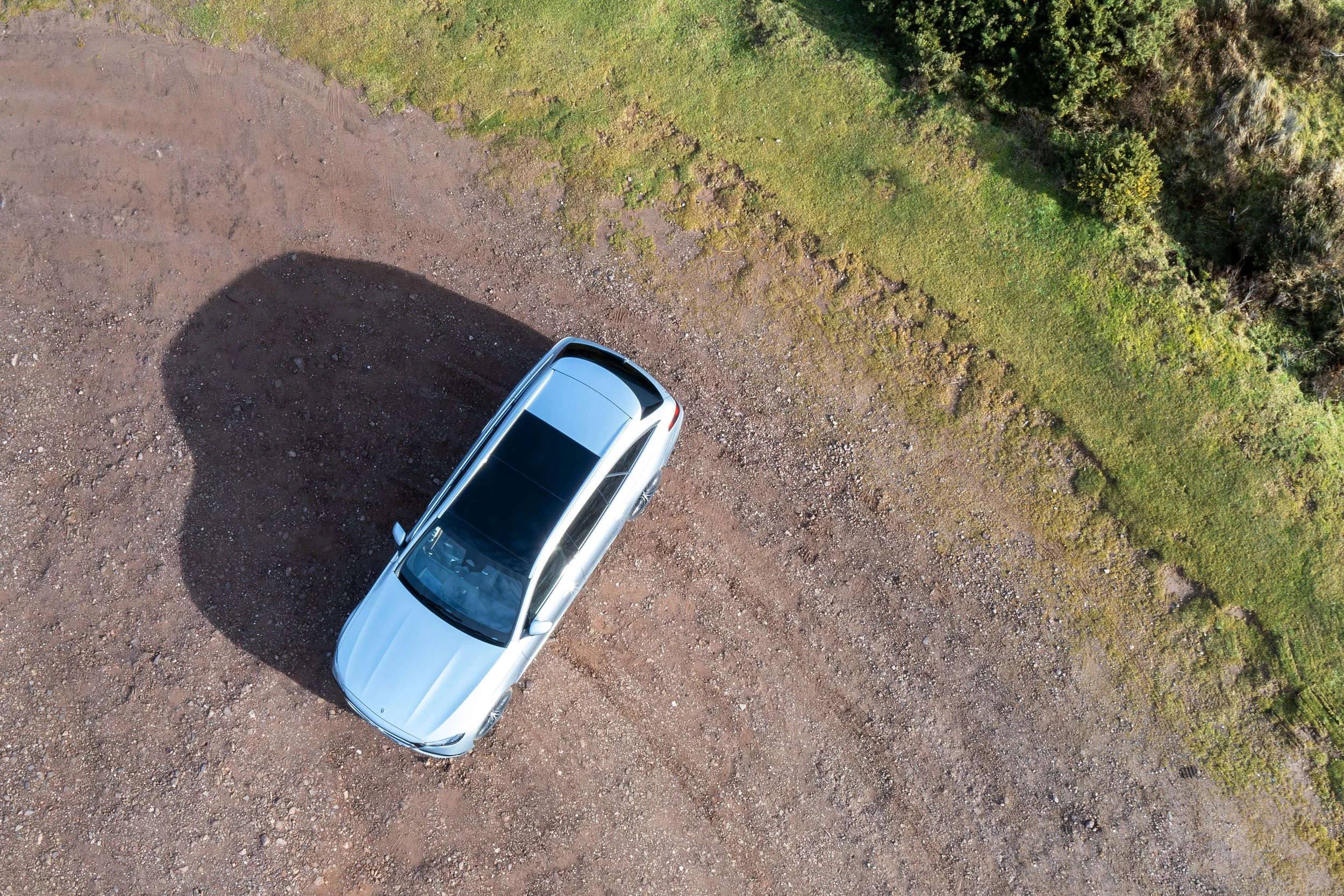 An overhead view of a silver car parked on a dirt area next to a grassy field with trees.