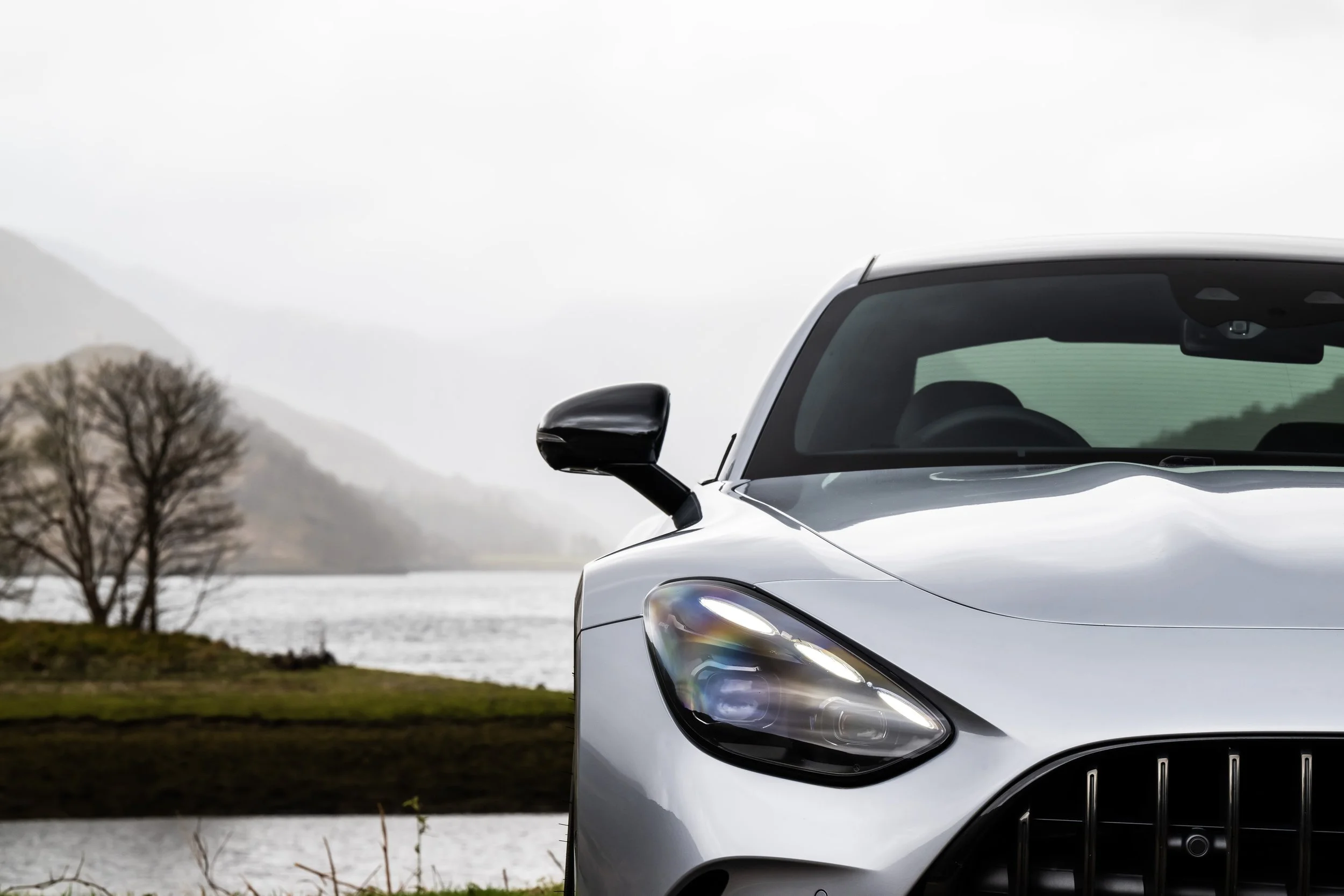 Close-up of the front of a silver sports car with a scenic lake and mountains in the background.