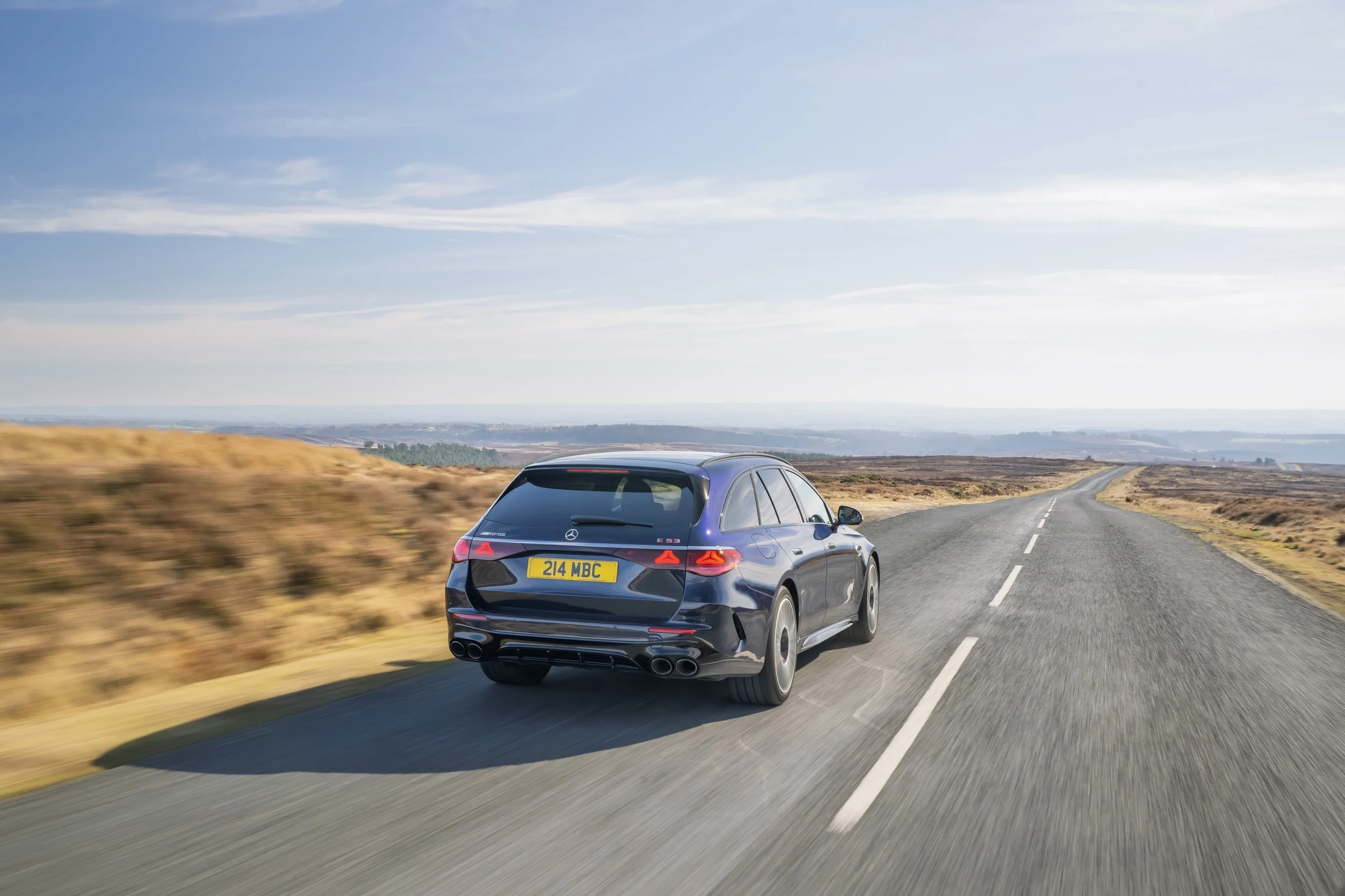 A black Mercedes-Benz E-Class wagon driving on a rural two-lane road surrounded by open fields under a partly cloudy sky.