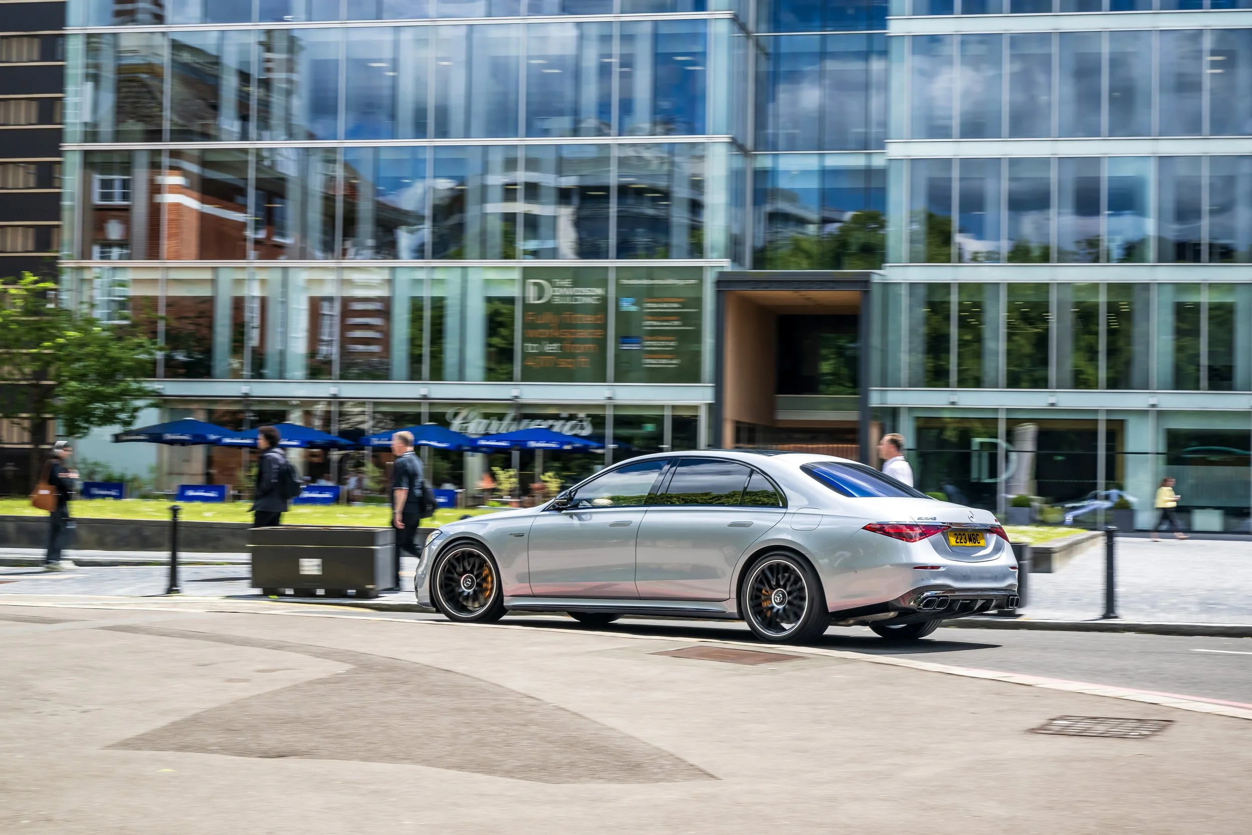 A silver luxury car parked on a city street in front of a glass building with pedestrians walking by.
