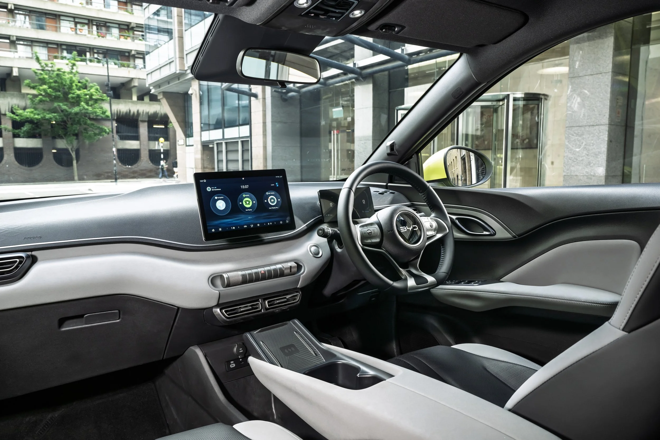 Interior of a modern electric vehicle with a digital dashboard, steering wheel, and touchscreen display, parked outside a glass building