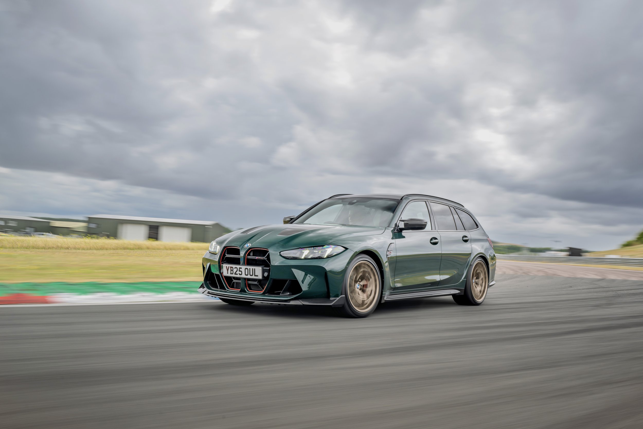 A green BMW station wagon racing on a track under cloudy skies, with blurred background and bronze-colored wheels.