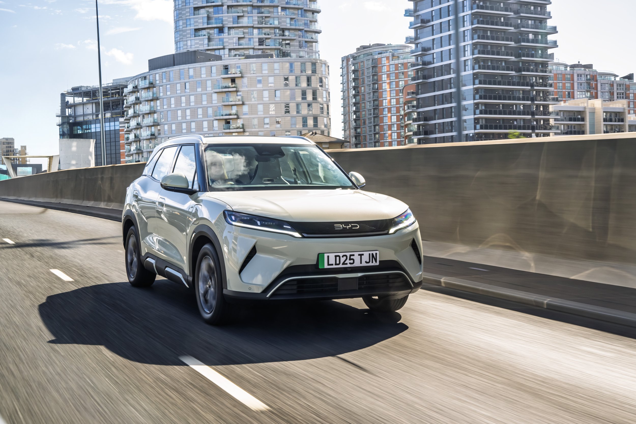 A white electric SUV with a UK license plate driving on a city highway with modern high-rise buildings in the background.
