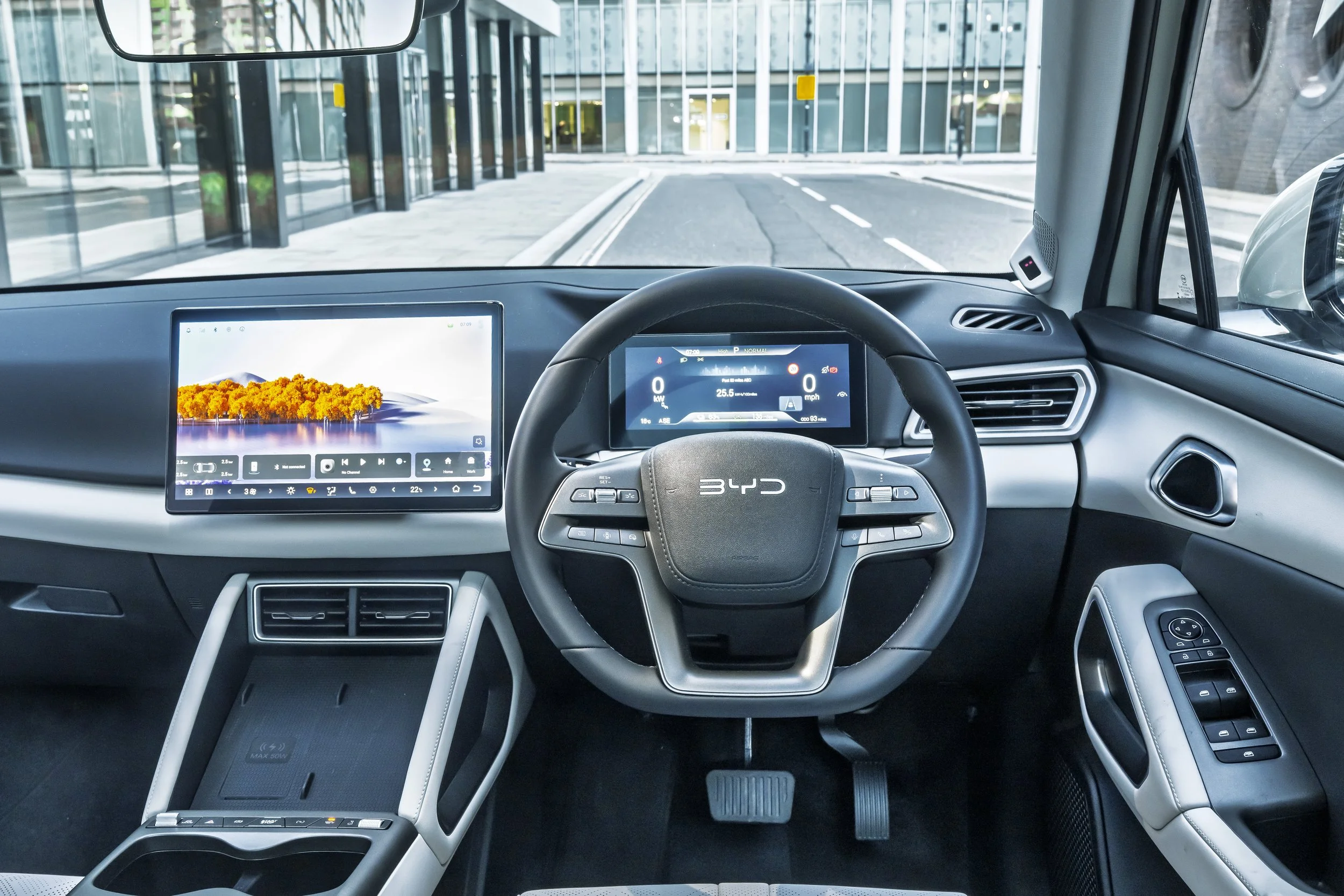 Interior of an electric BYD vehicle showing digital dashboard, steering wheel, and infotainment screens, with a city street view through the windshield.