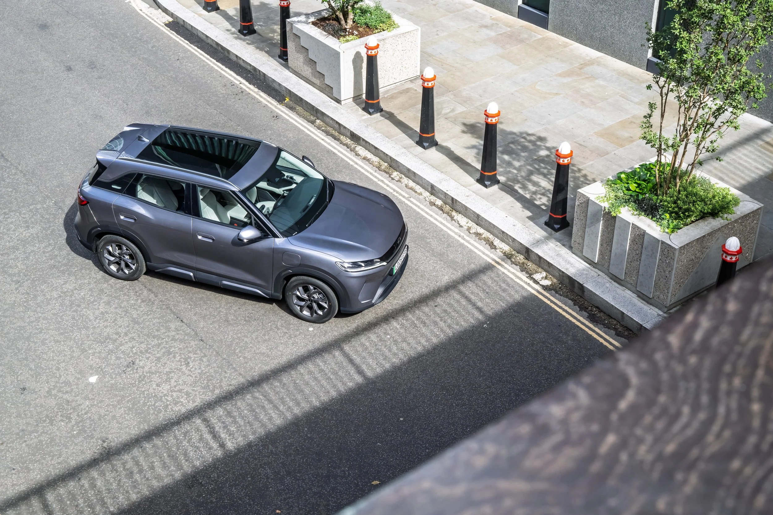 A gray SUV parked on the side of a city street next to a sidewalk with large planters containing small trees and plants, with black bollards and a building in the background.