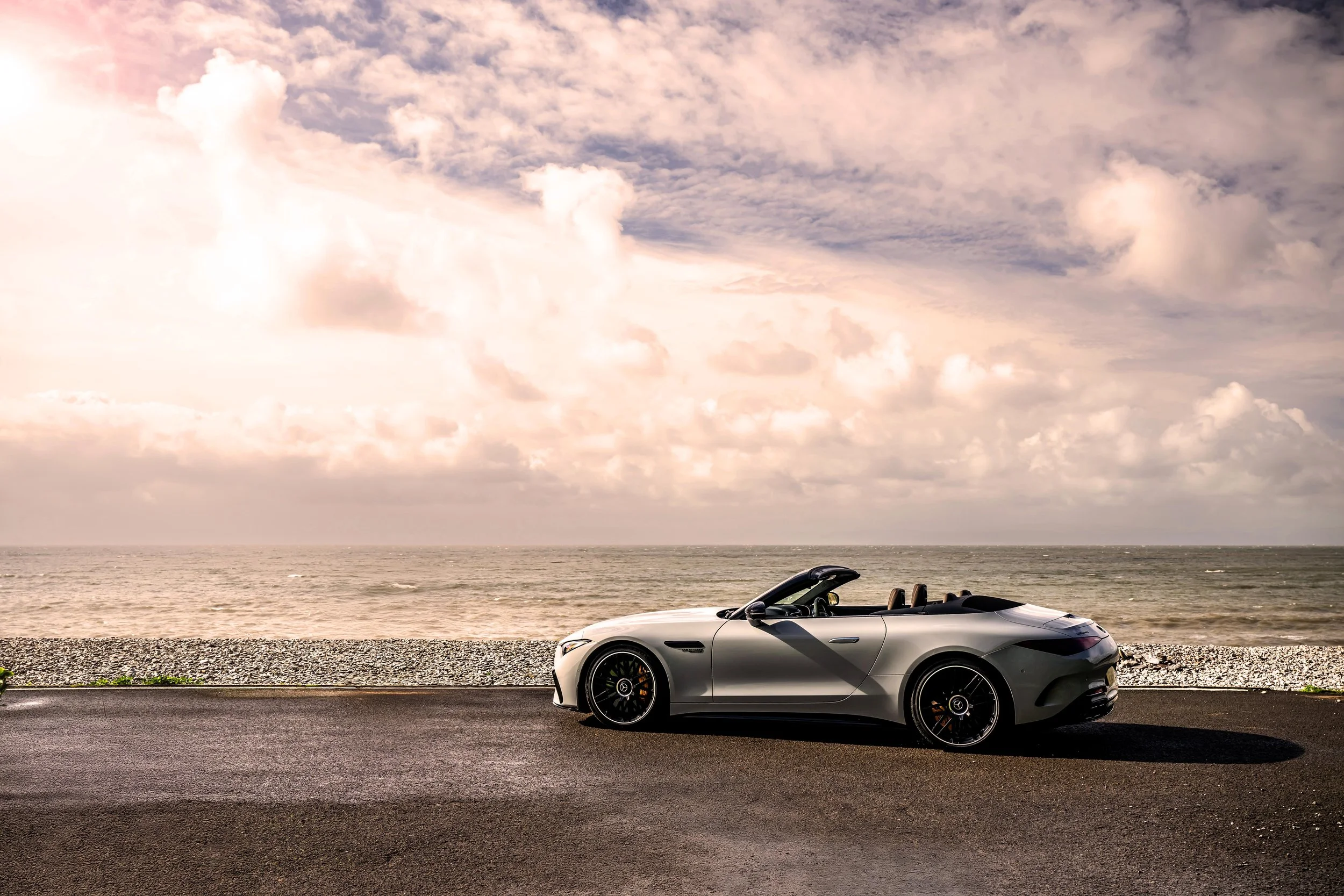 A silver convertible sports car parked on a paved road beside a gravel path, with the ocean and a cloudy sky in the background.