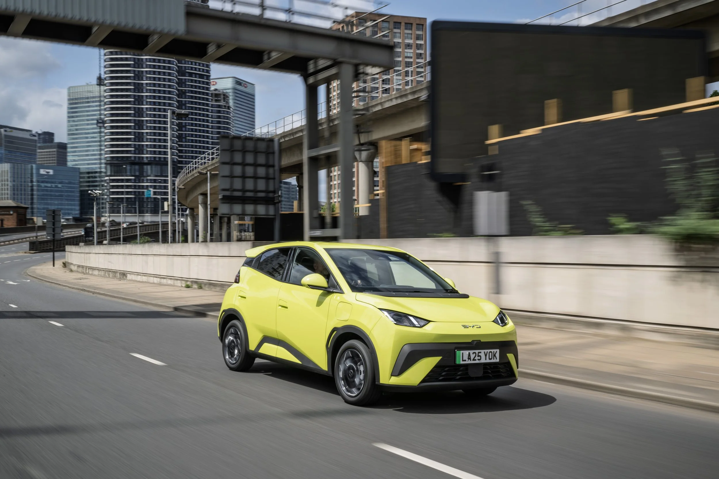 A bright yellow compact electric car driving on an urban highway with city skyscrapers and overpasses in the background.