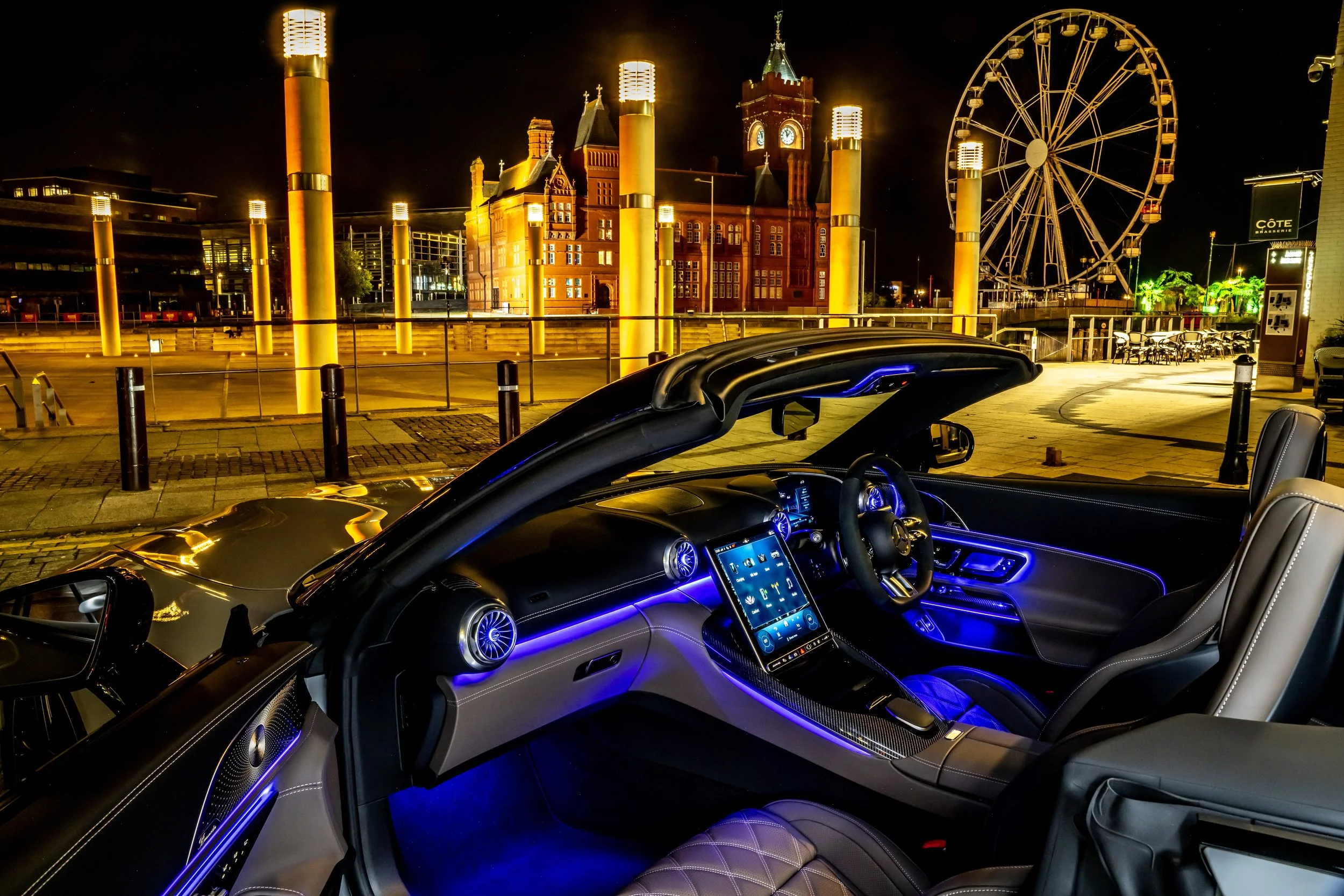 The interior of a luxurious convertible car with illuminated blue accents, parked near a brightly lit cityscape at night featuring a historic red brick building, a large Ferris wheel, and modern buildings in the background.