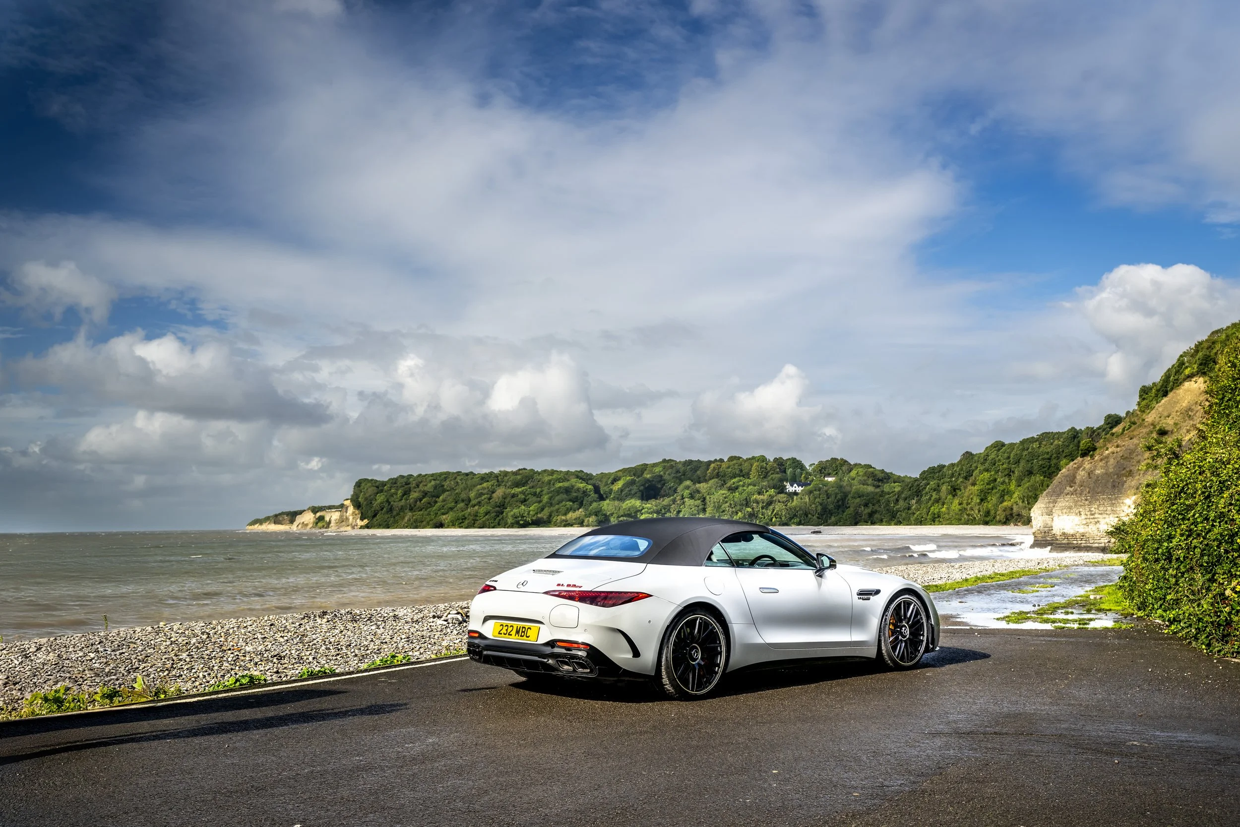 A white convertible sports car with black wheels parked on a paved road near a beach with water, sandy cliffs, and green hills under a partly cloudy sky.