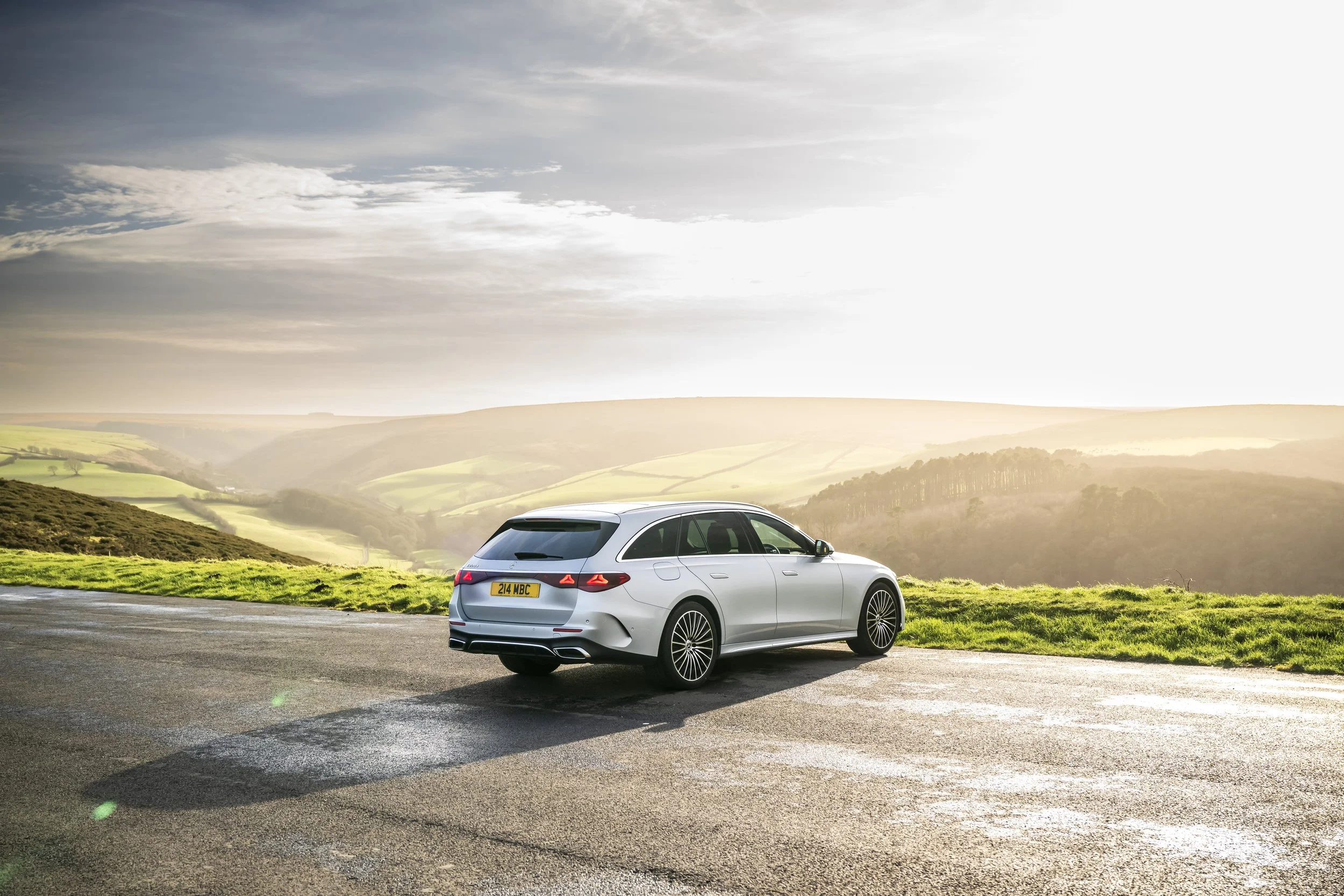 A silver station wagon parked on a rural road with rolling green hills in the background during sunset or sunrise.