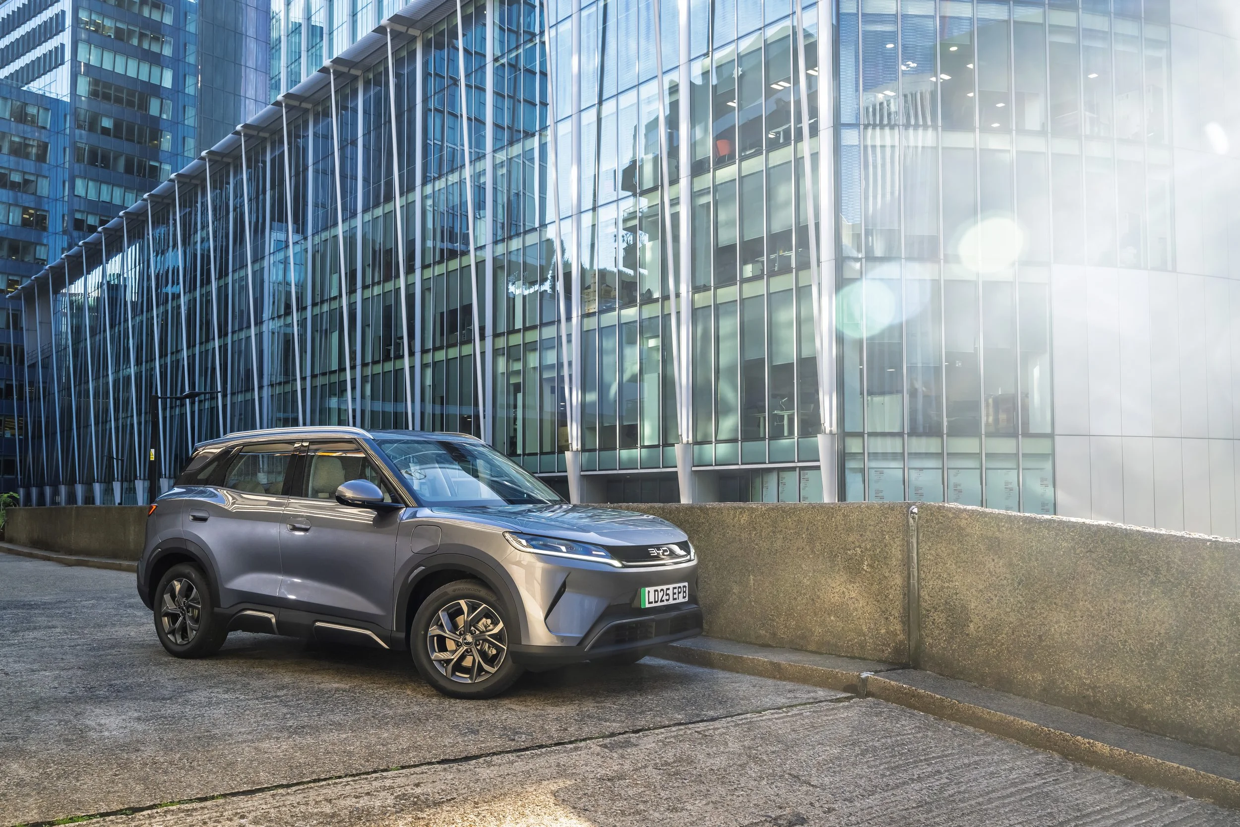 A silver electric car parked outside a modern glass building with metal structural elements, under bright sunlight.