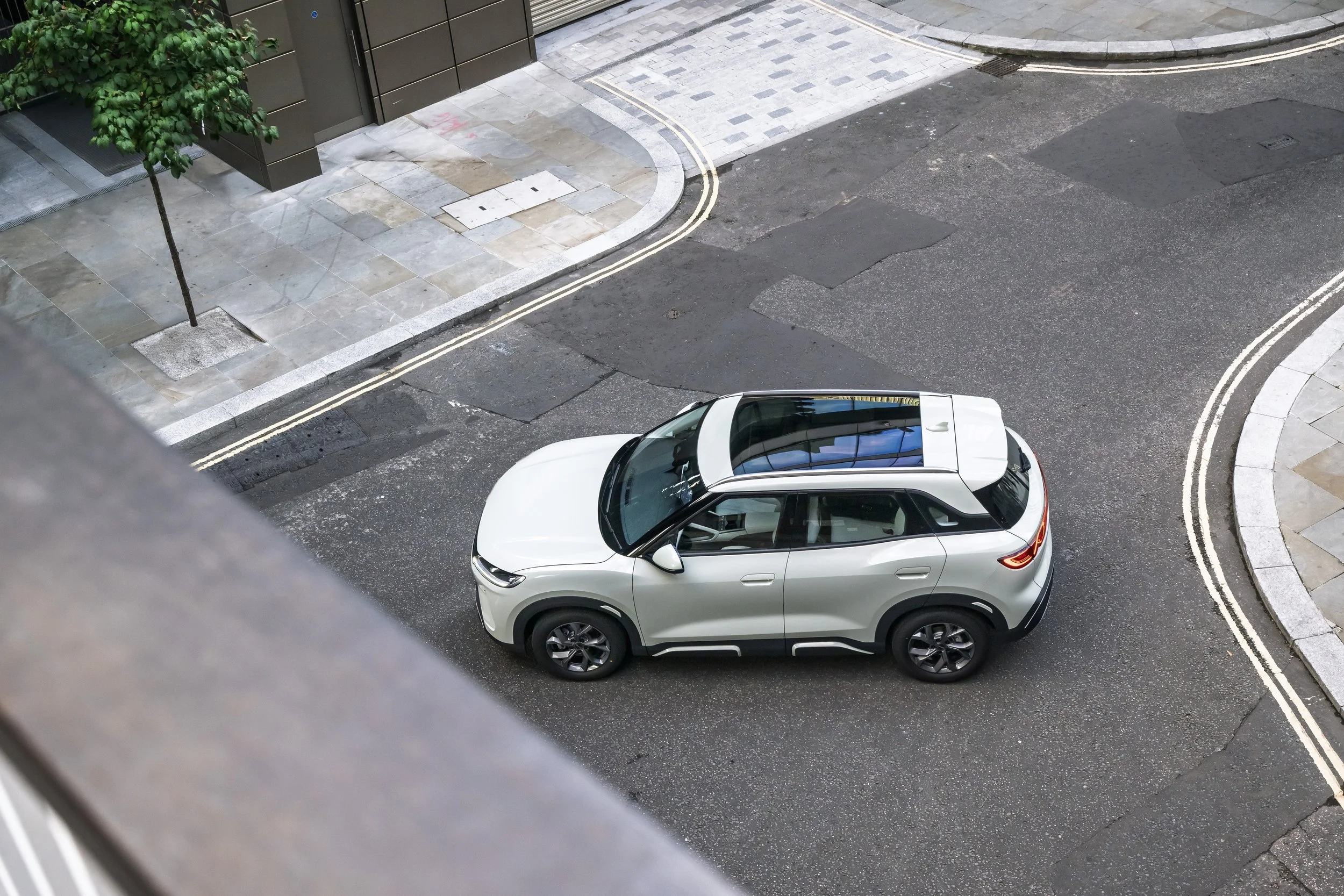 A white SUV parked on a city street with a sidewalk and a tree visible, seen from an elevated perspective.
