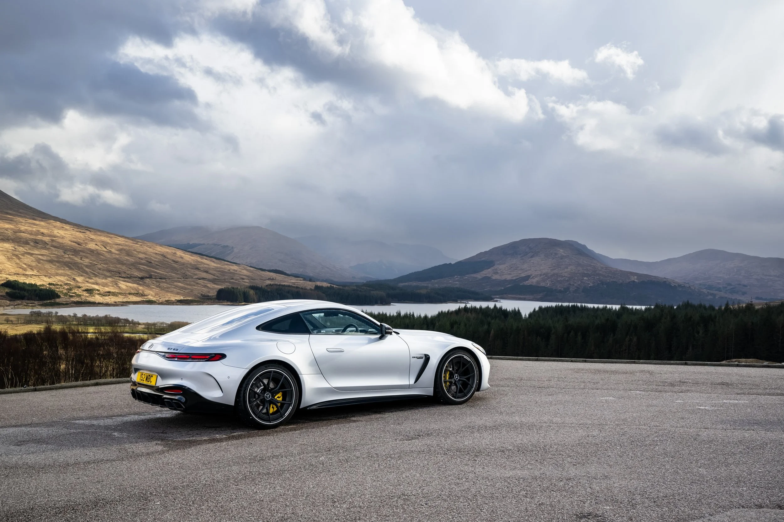 A white luxury sports car parked on an empty road with mountains, a lake, and cloudy sky in the background.