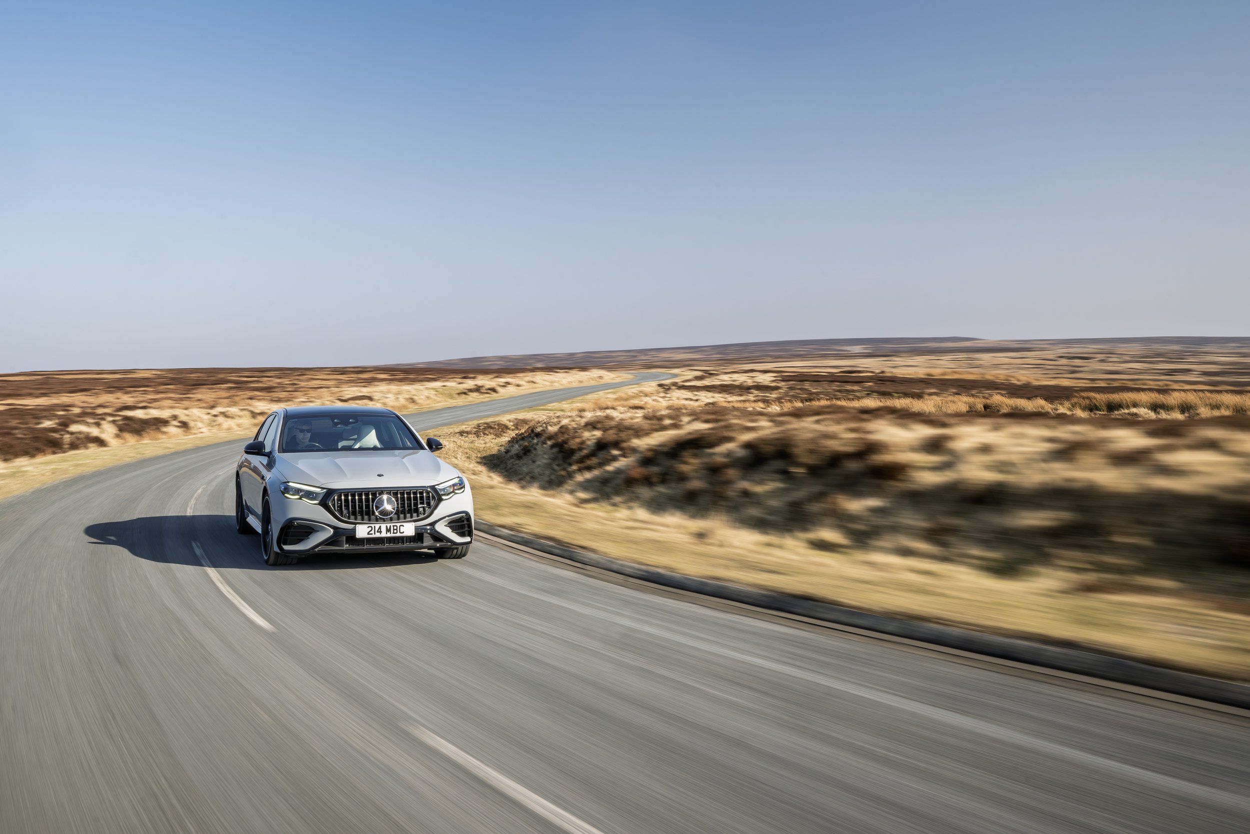 A silver sedan driving on a winding desert road with dry grass and hills in the background under a clear blue sky.