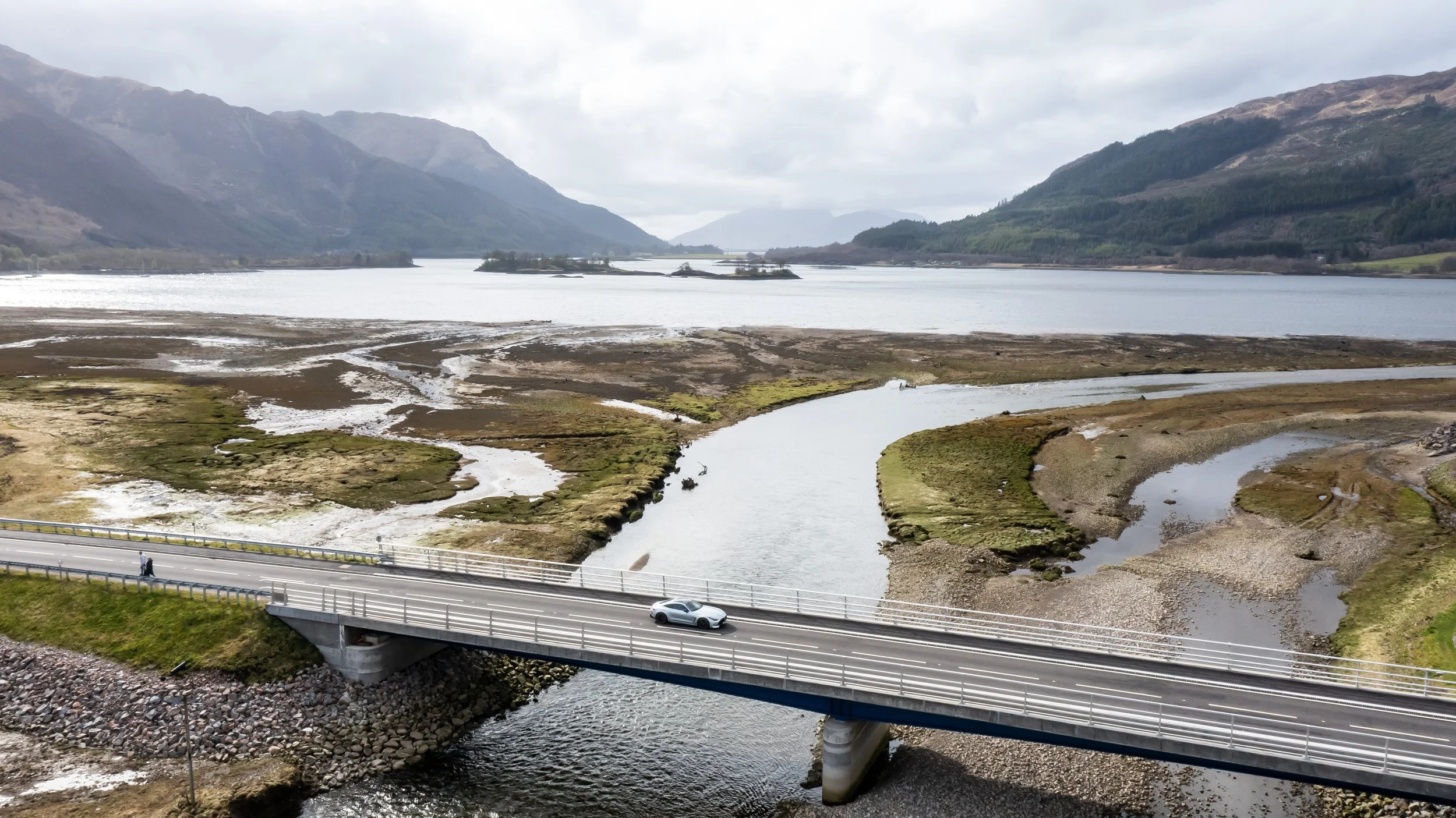 A bridge crossing over a river with a car on it, surrounded by wetlands, small streams, and mountains in the background under a cloudy sky.