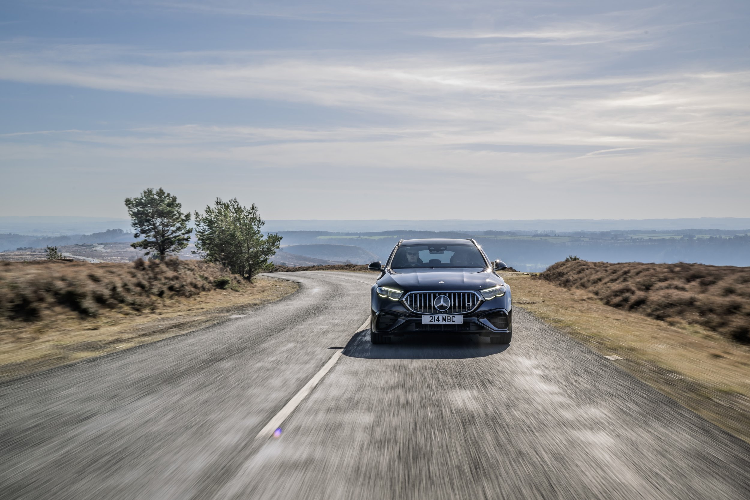 A black Mercedes-Benz car driving on a winding road in a rural landscape with trees and hills in the background under a partly cloudy sky.