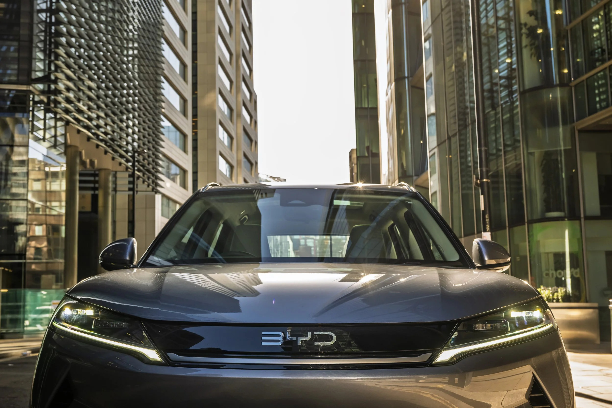 Black BYD electric car parked on a city street with tall glass buildings on either side.