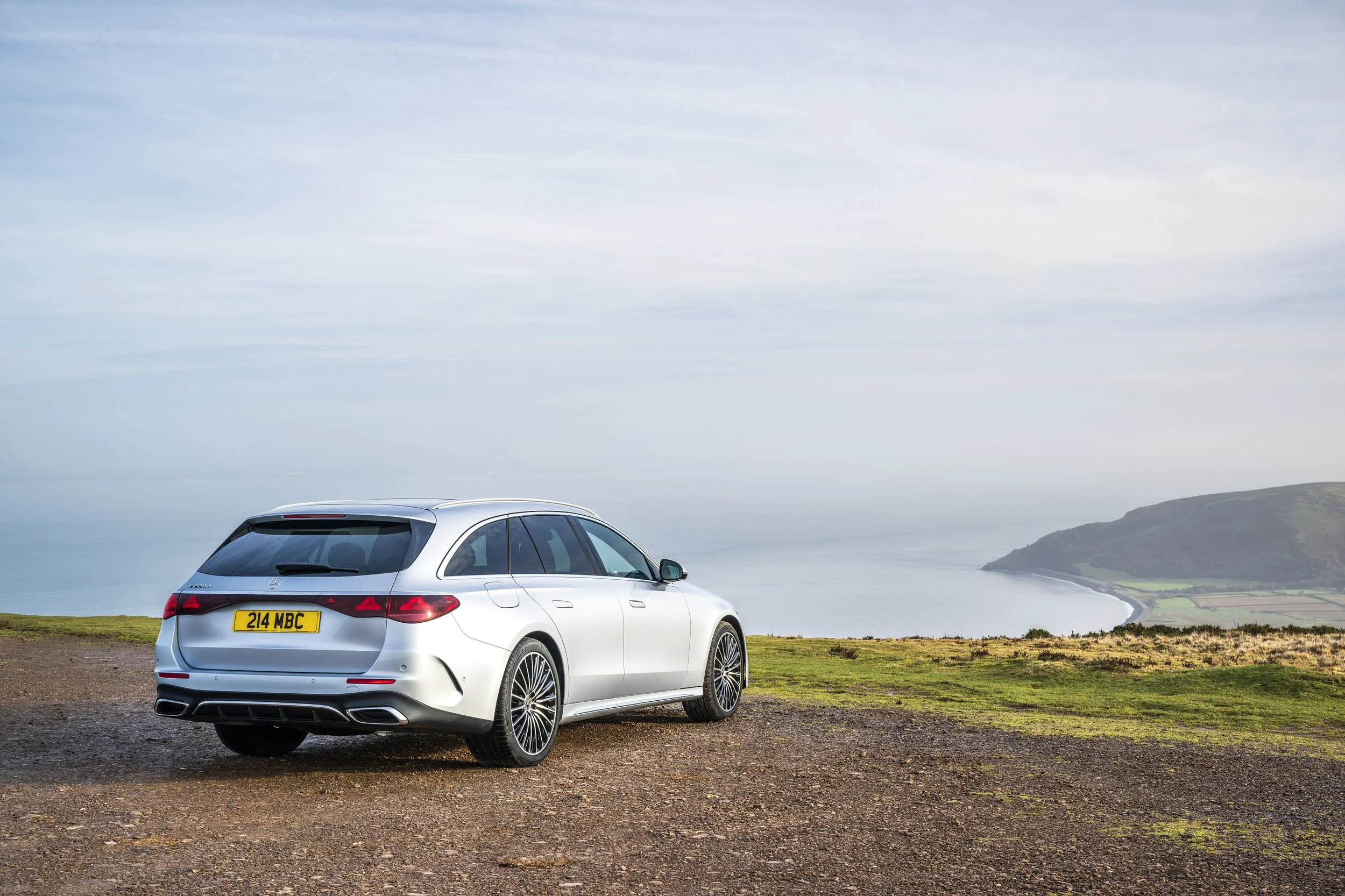 A white Mercedes-Benz station wagon parked on a grassy cliff overlooking the ocean with a cloudy sky.