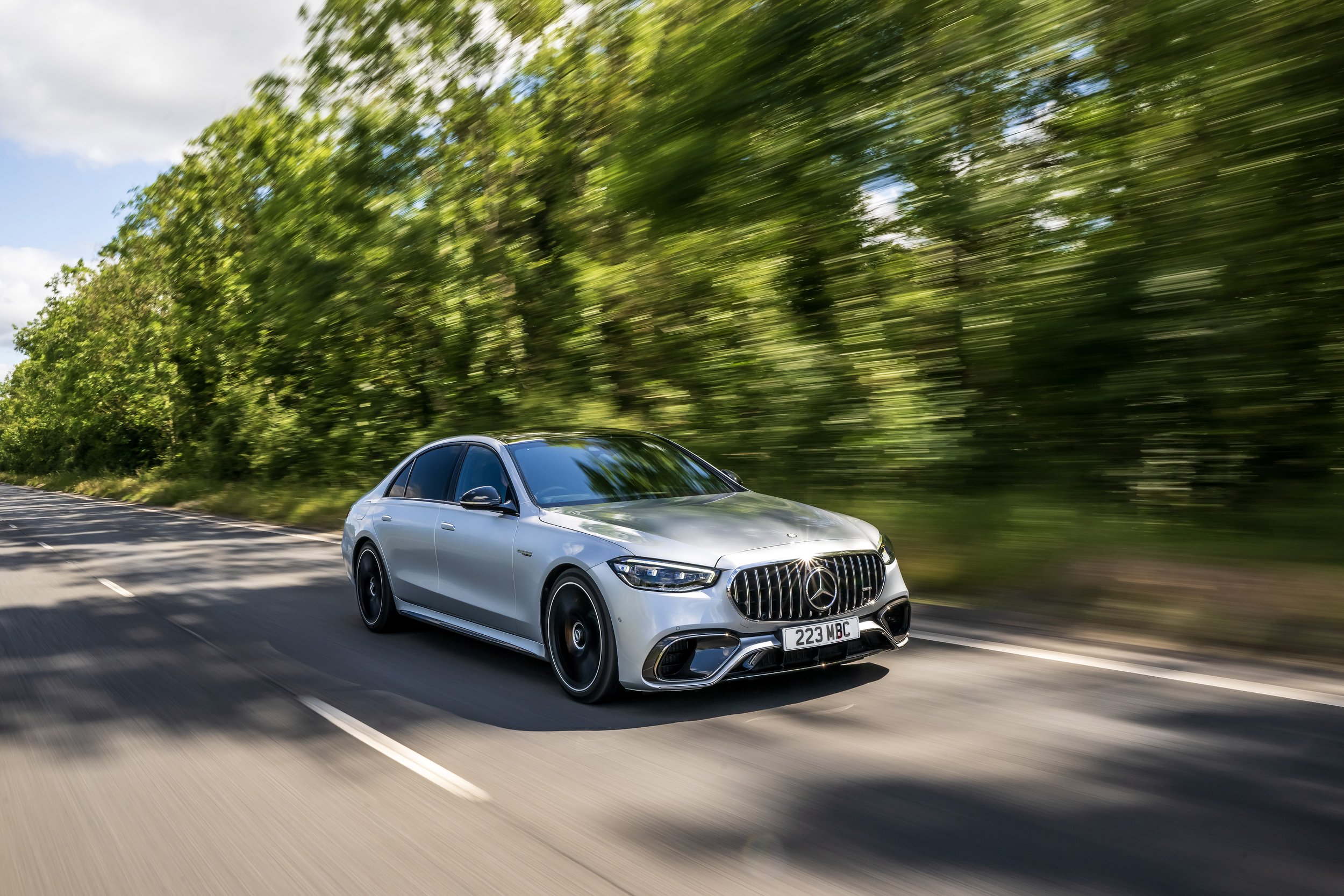 A silver Mercedes-Benz luxury sedan driving on a countryside road with green trees and blue sky in the background.