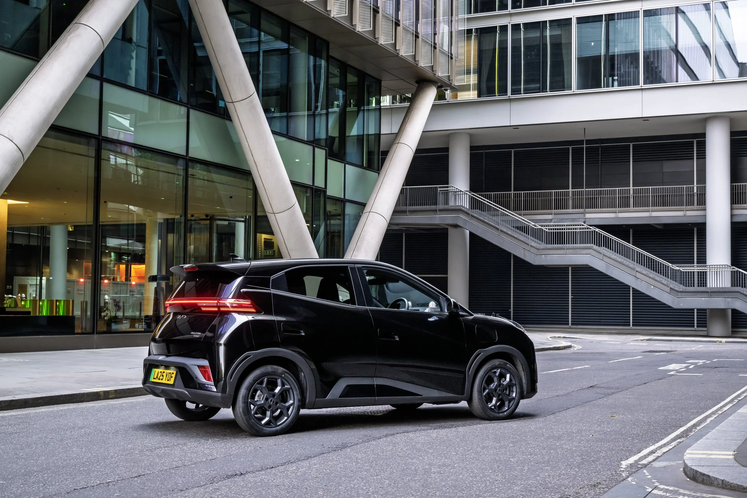 A black electric car parked on a city street near a modern glass building with structural supports and exterior staircases.