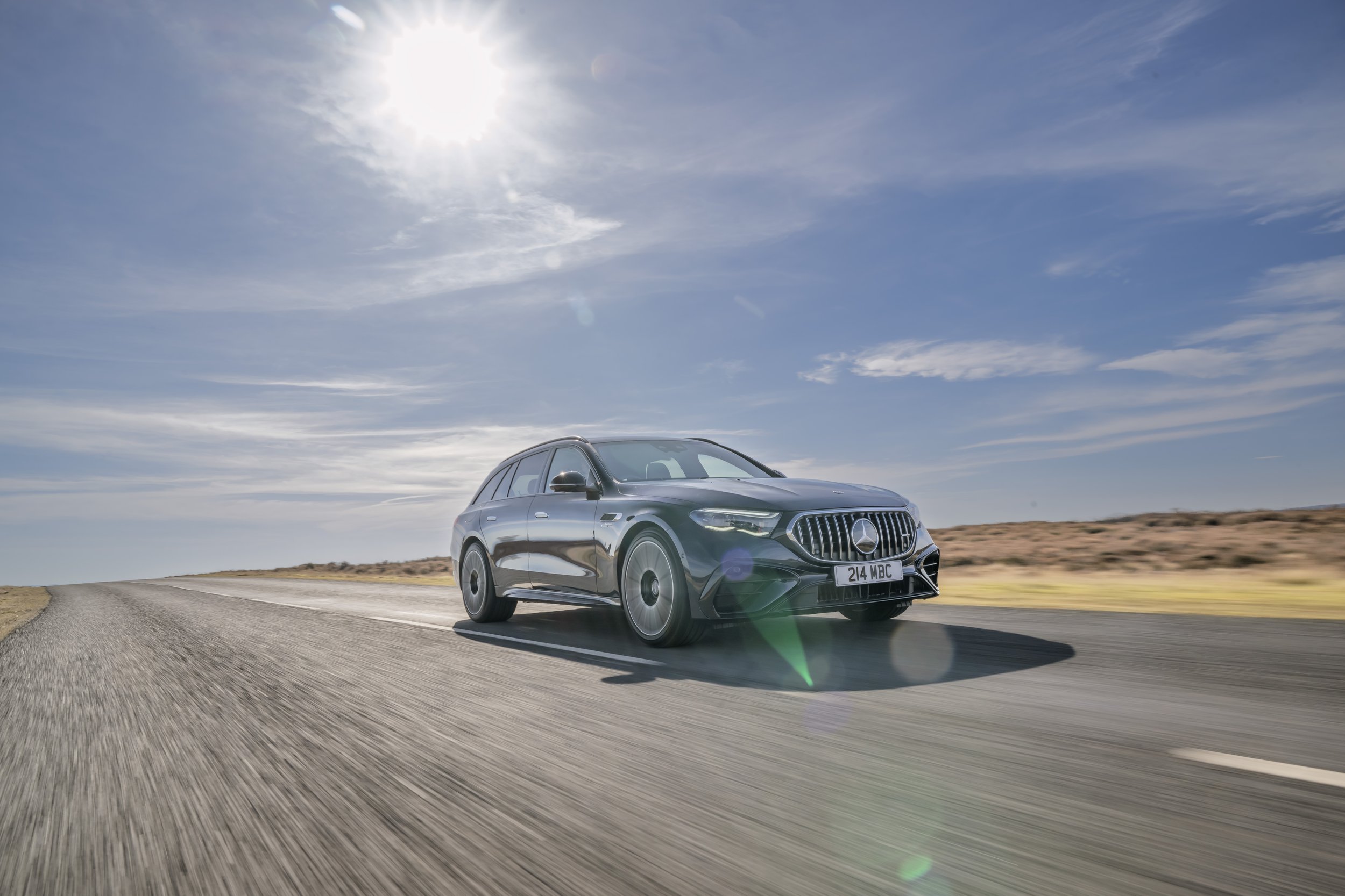 A black Mercedes-Benz station wagon driving on a paved road in a desert landscape under a bright blue sky with clouds and the sun shining.