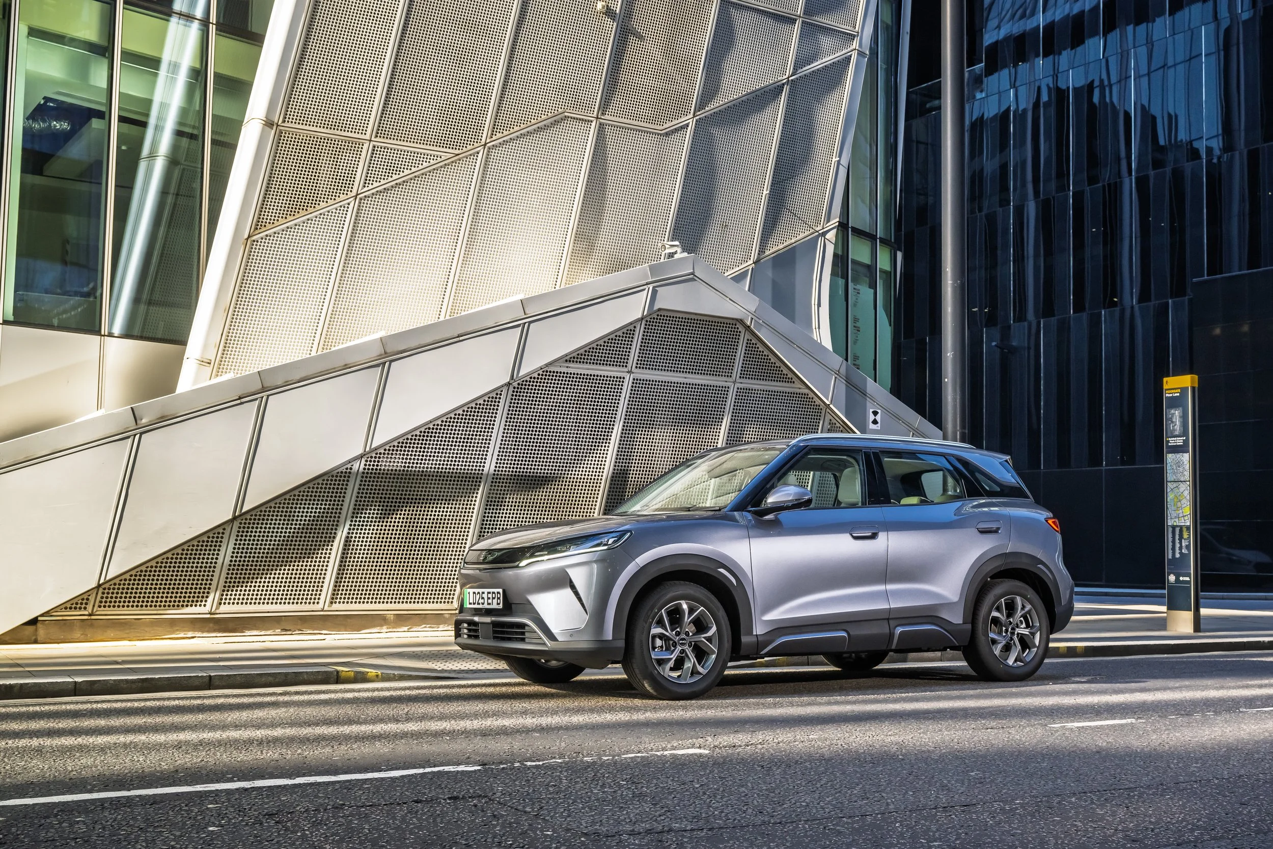 A silver compact SUV parked on a city street in front of modern glass and steel buildings, with a bus stop sign on the sidewalk.