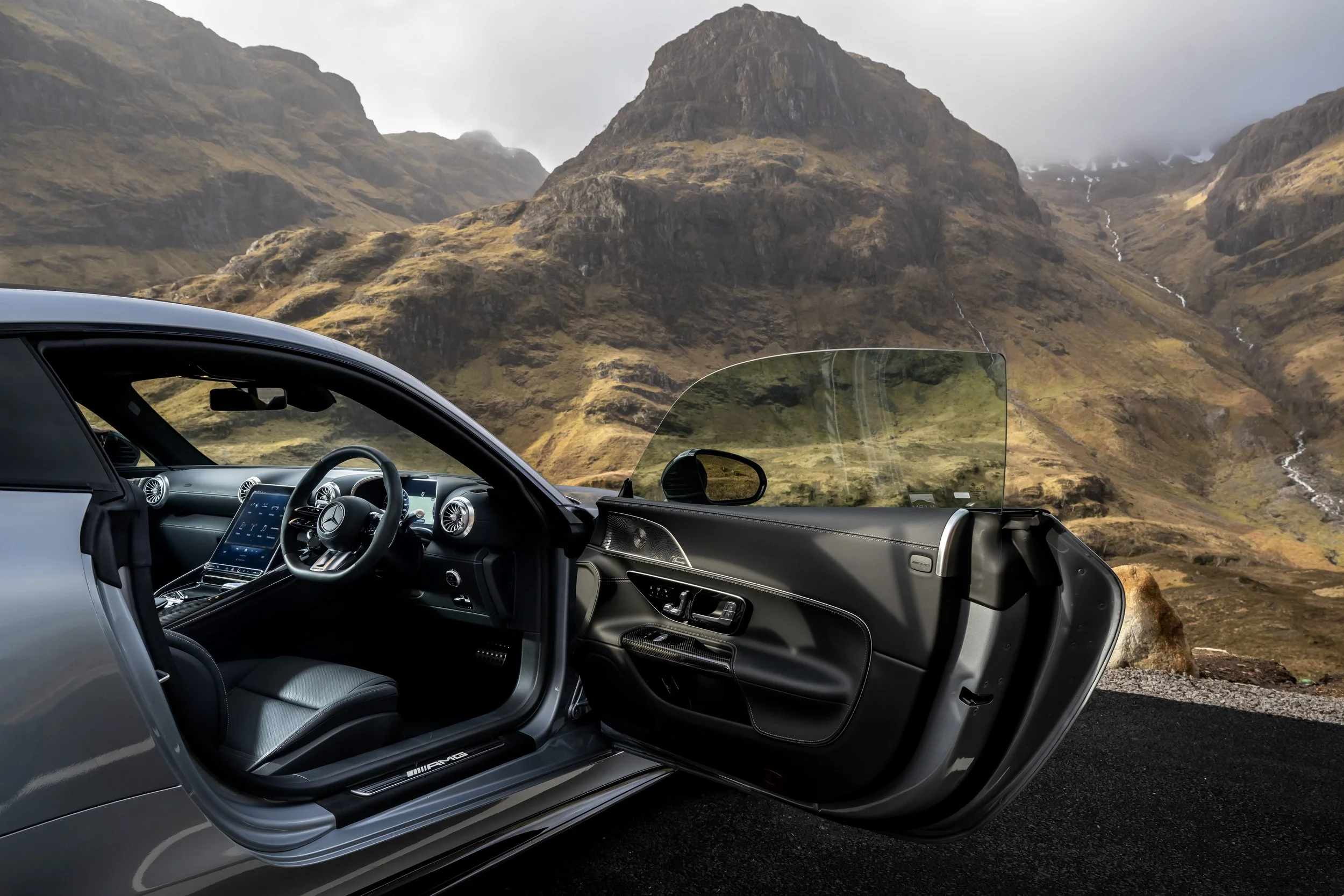 Interior of a silver luxury car with the driver's side door open, set against a mountainous landscape with steep grassy hills and streams.