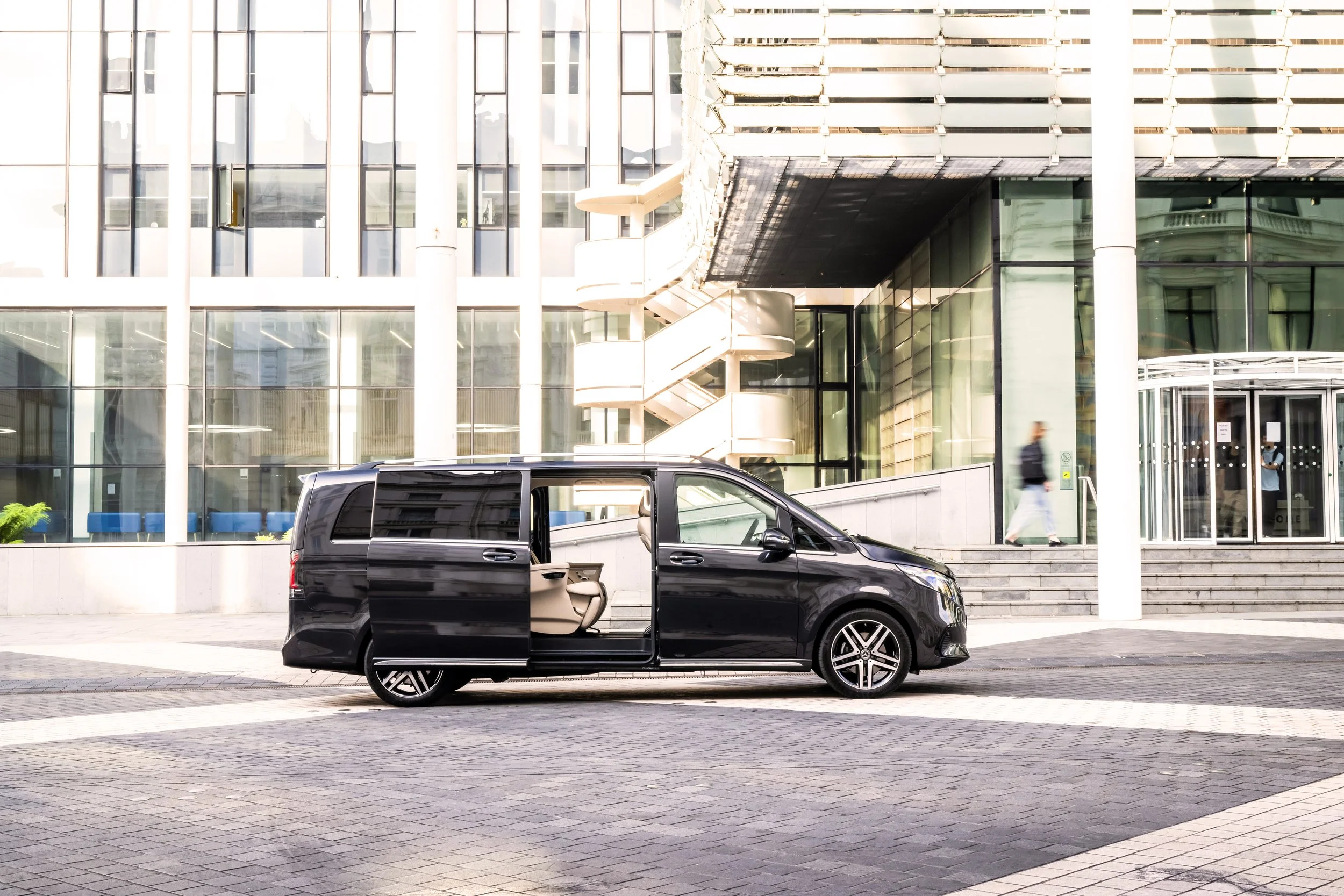 A black minivan parked in front of a modern office building with glass windows and a staircase outside. One sliding door of the van is open, revealing beige interior seats. A person is walking up the steps toward the entrance.