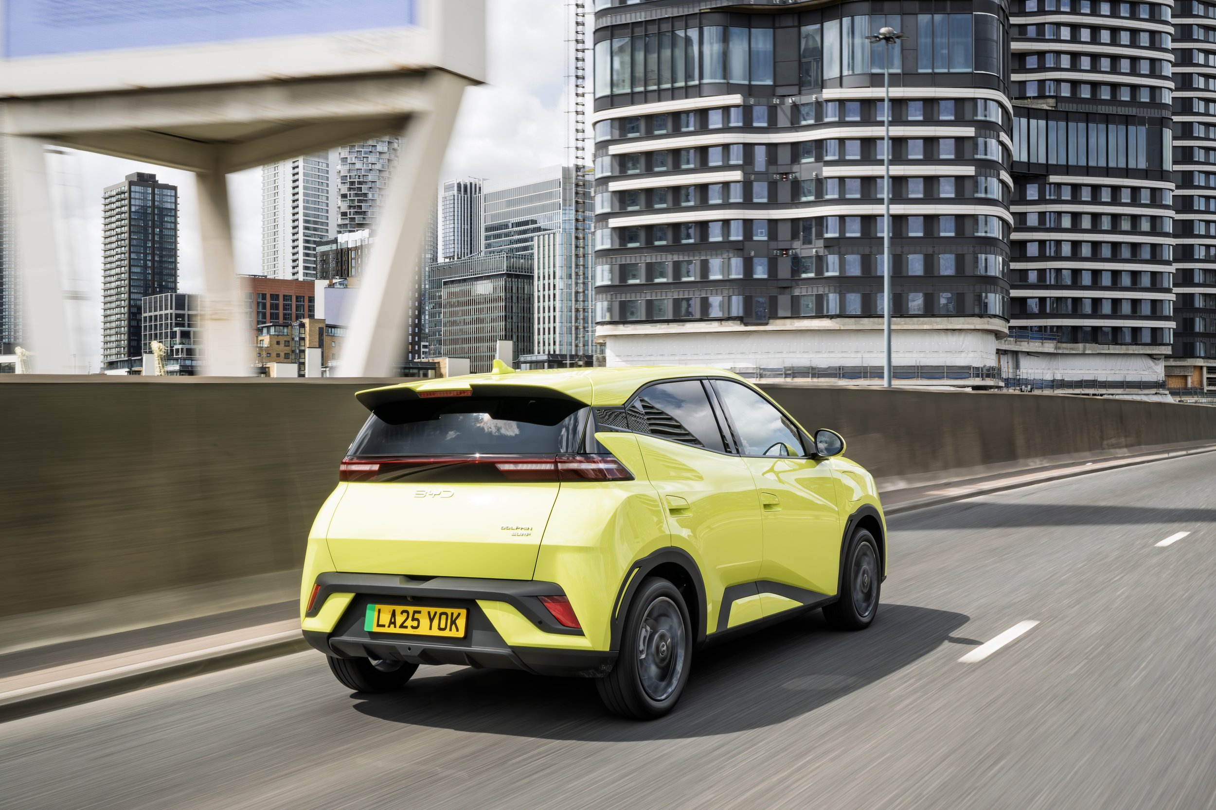 A yellow compact electric vehicle driving on a city highway with tall modern skyscrapers in the background.