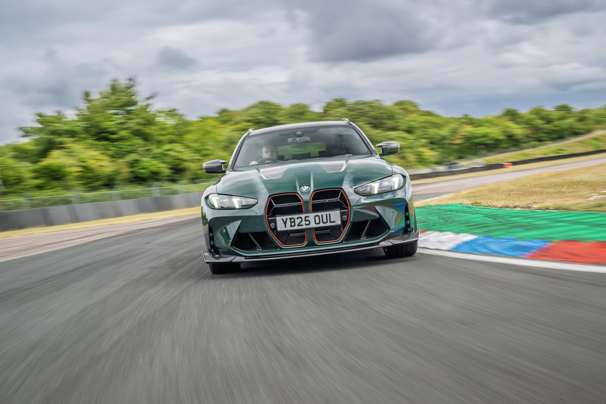 A green BMW car with a black and red front grille driving on a race track during cloudy weather.