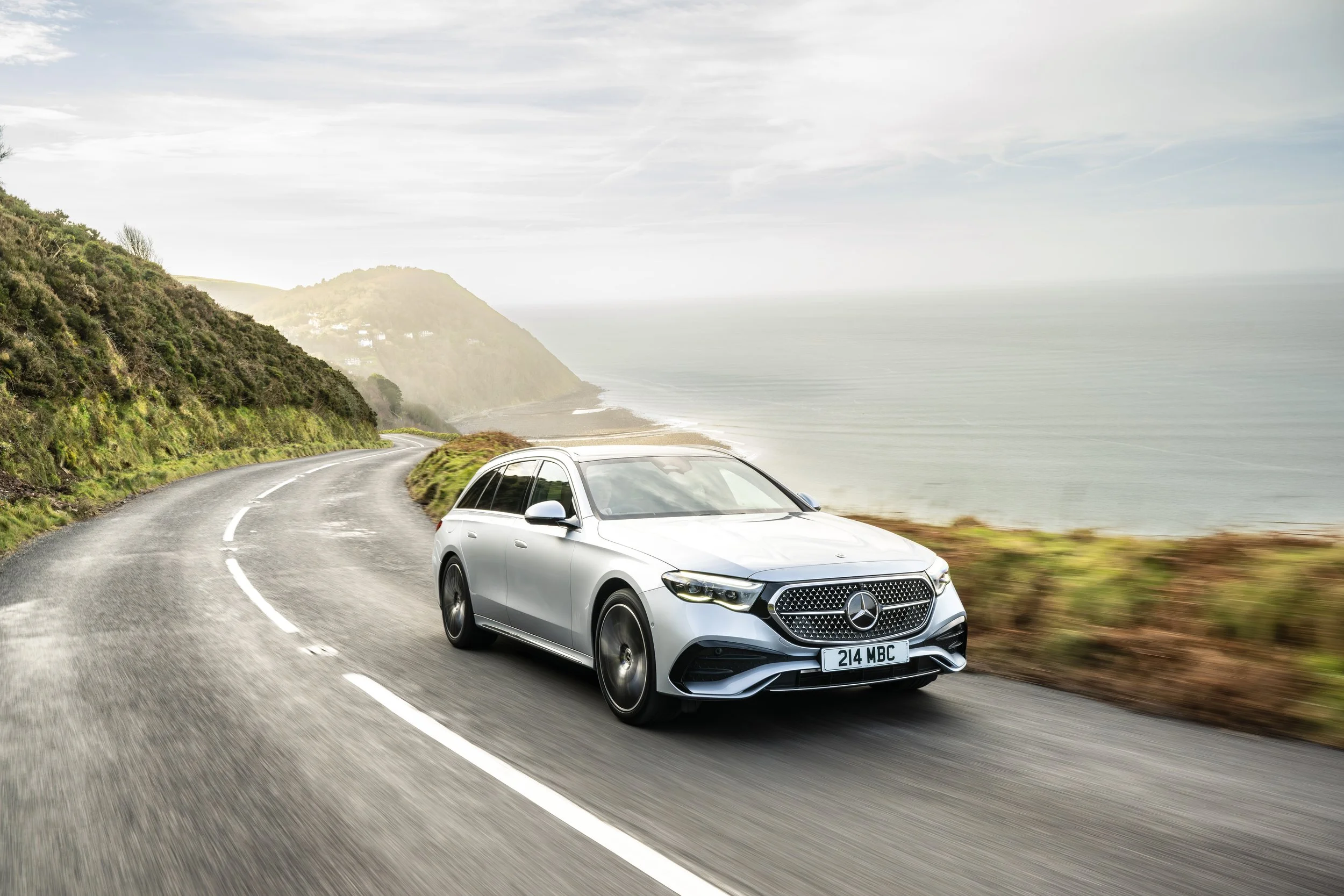 Silver Mercedes-Benz car driving on a winding coastal road with green hills and ocean in the background.