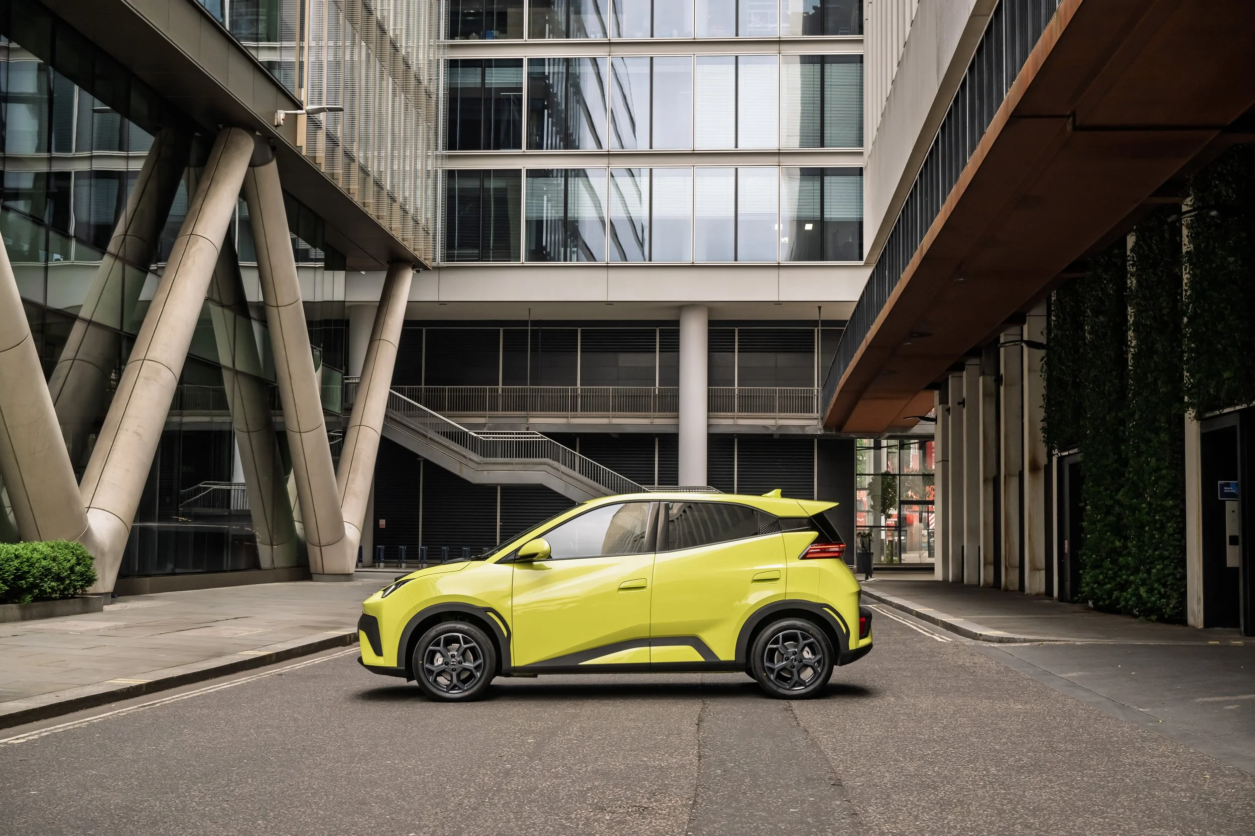 A yellow compact electric vehicle parked on city street in front of modern office building with glass windows and architectural supports.