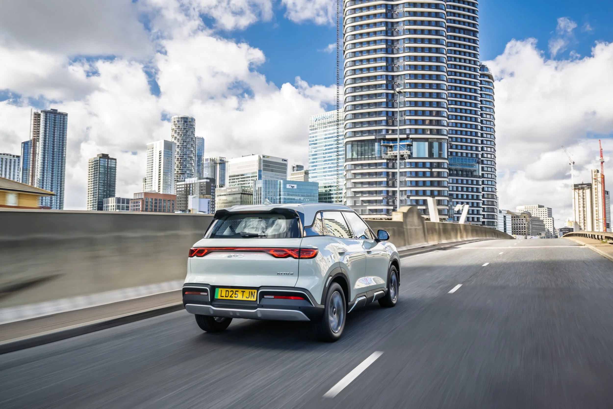 A white electric SUV driving on a city highway with tall modern skyscrapers in the background under a partly cloudy sky.