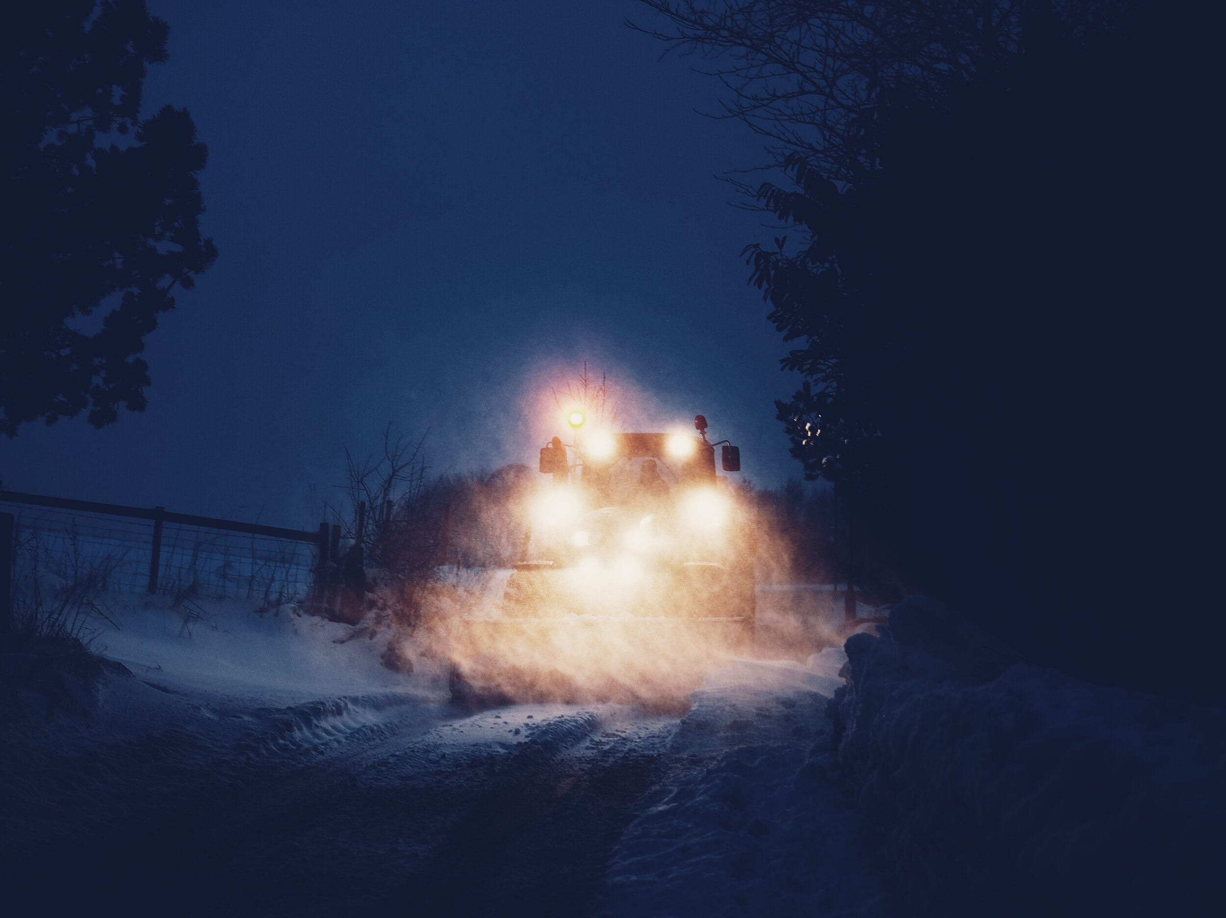 A snow-covered street at night with a snowplow or street cleaning vehicle with bright headlights clearing snow, surrounded by snowbanks and dark trees.