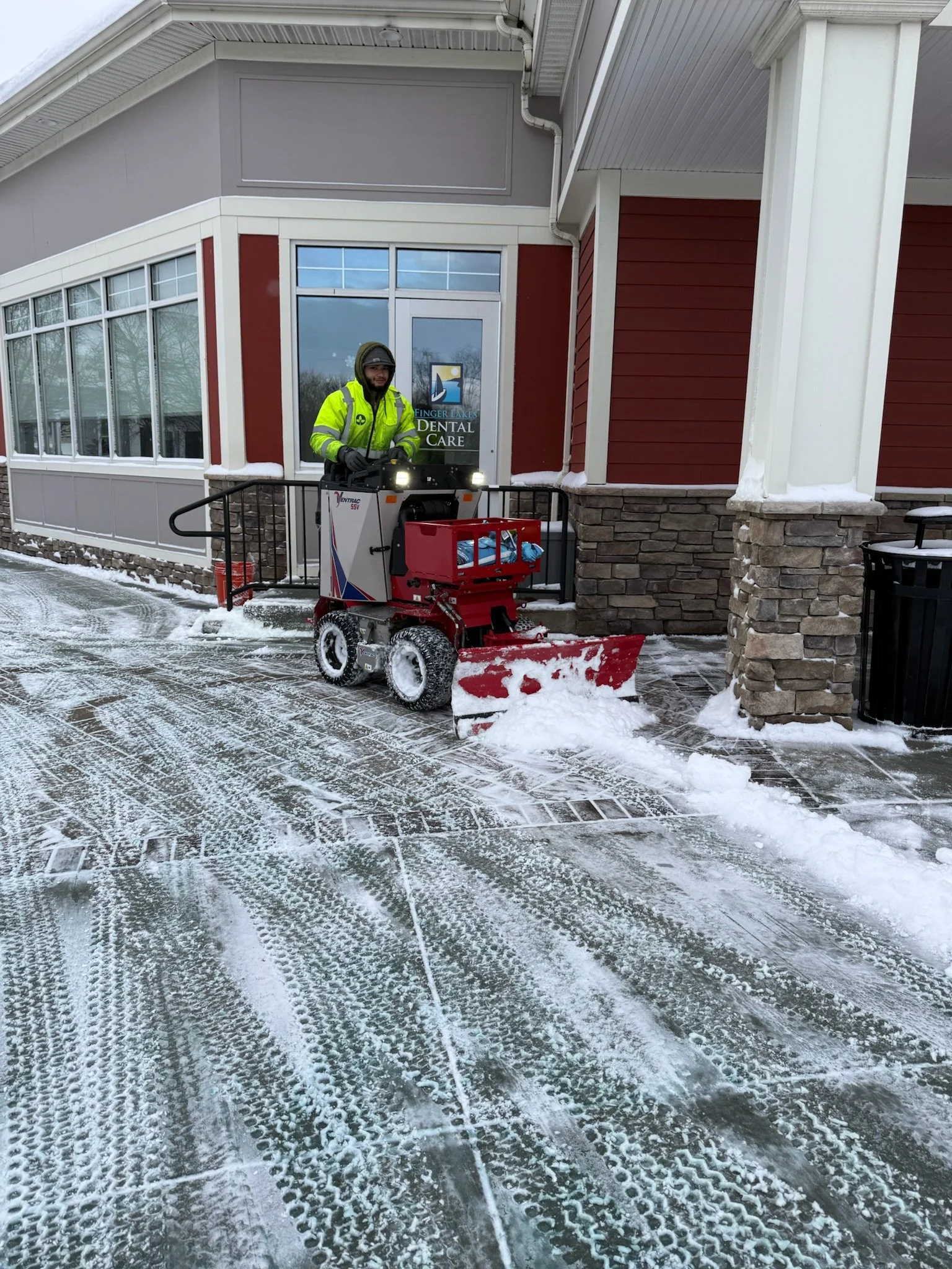 A man in a bright yellow jacket operating a snowplow on a snow-covered sidewalk in front of a building with red and gray siding, glass doors, and stone accents.