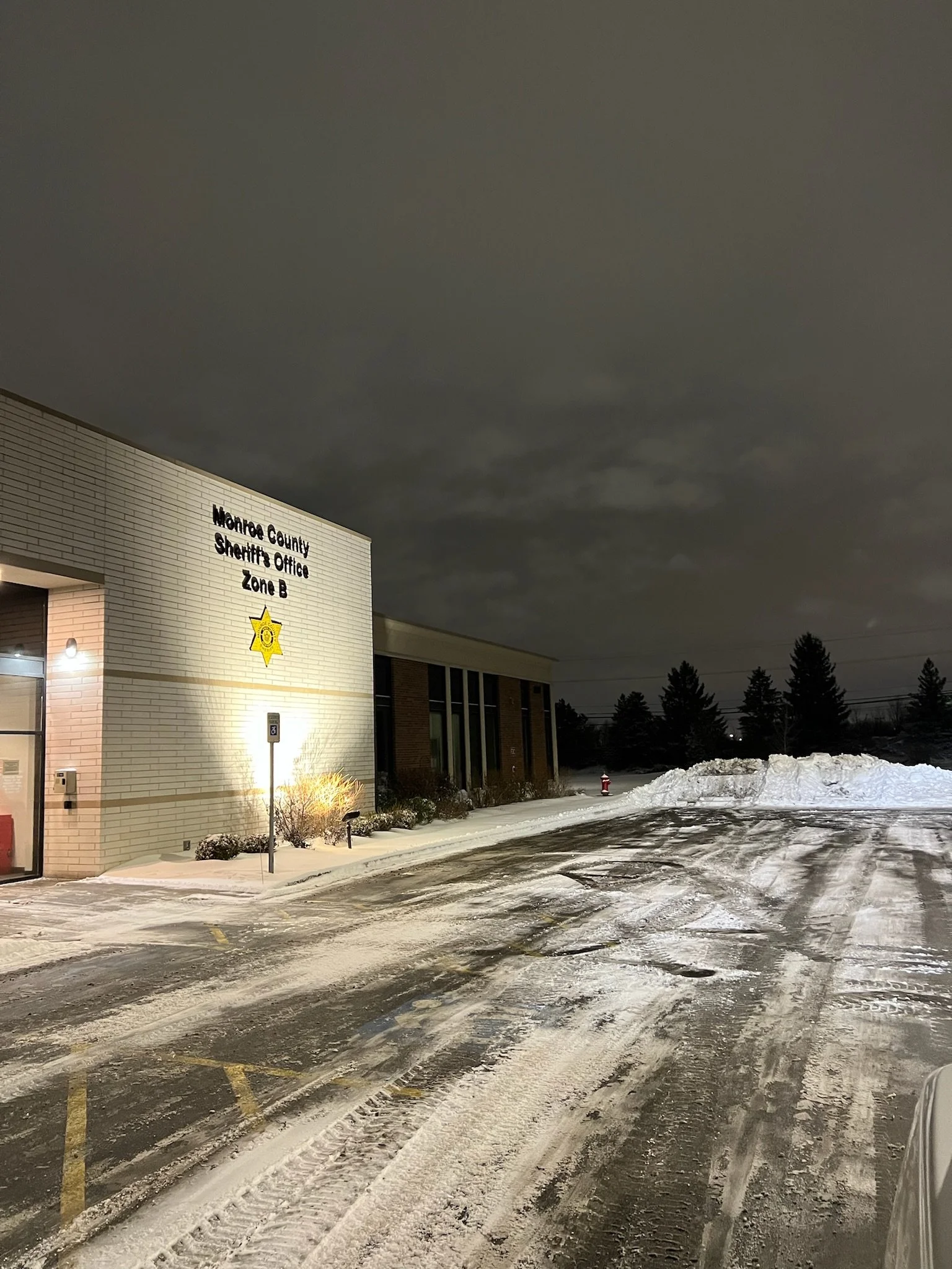 Nighttime scene outside a Monroe County Sheriff's Office building with snow on the parking lot and snow piles on the side and trees in the background.