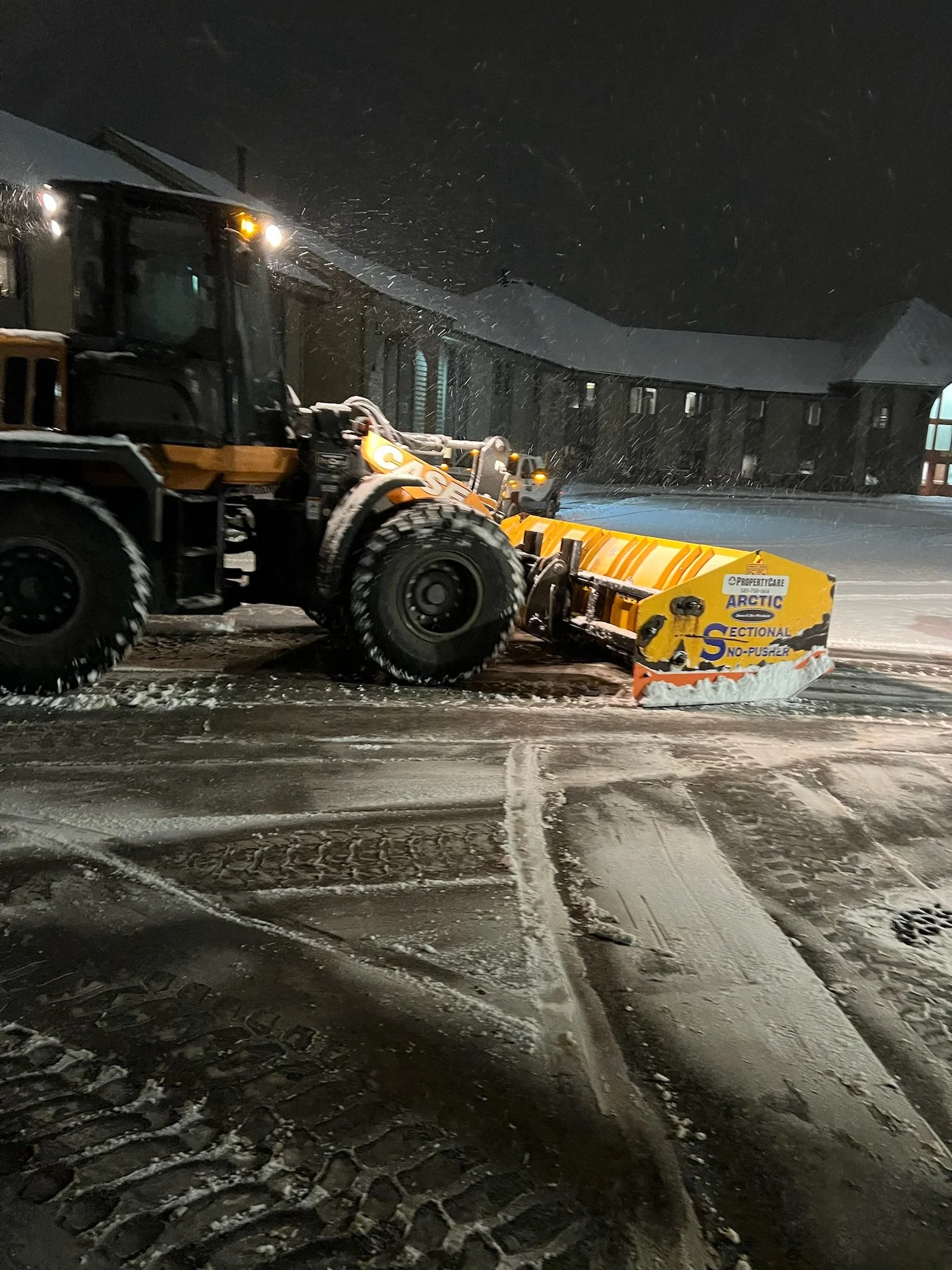 A snowplow clearing an icy, snow-covered street at night, with snow falling around.