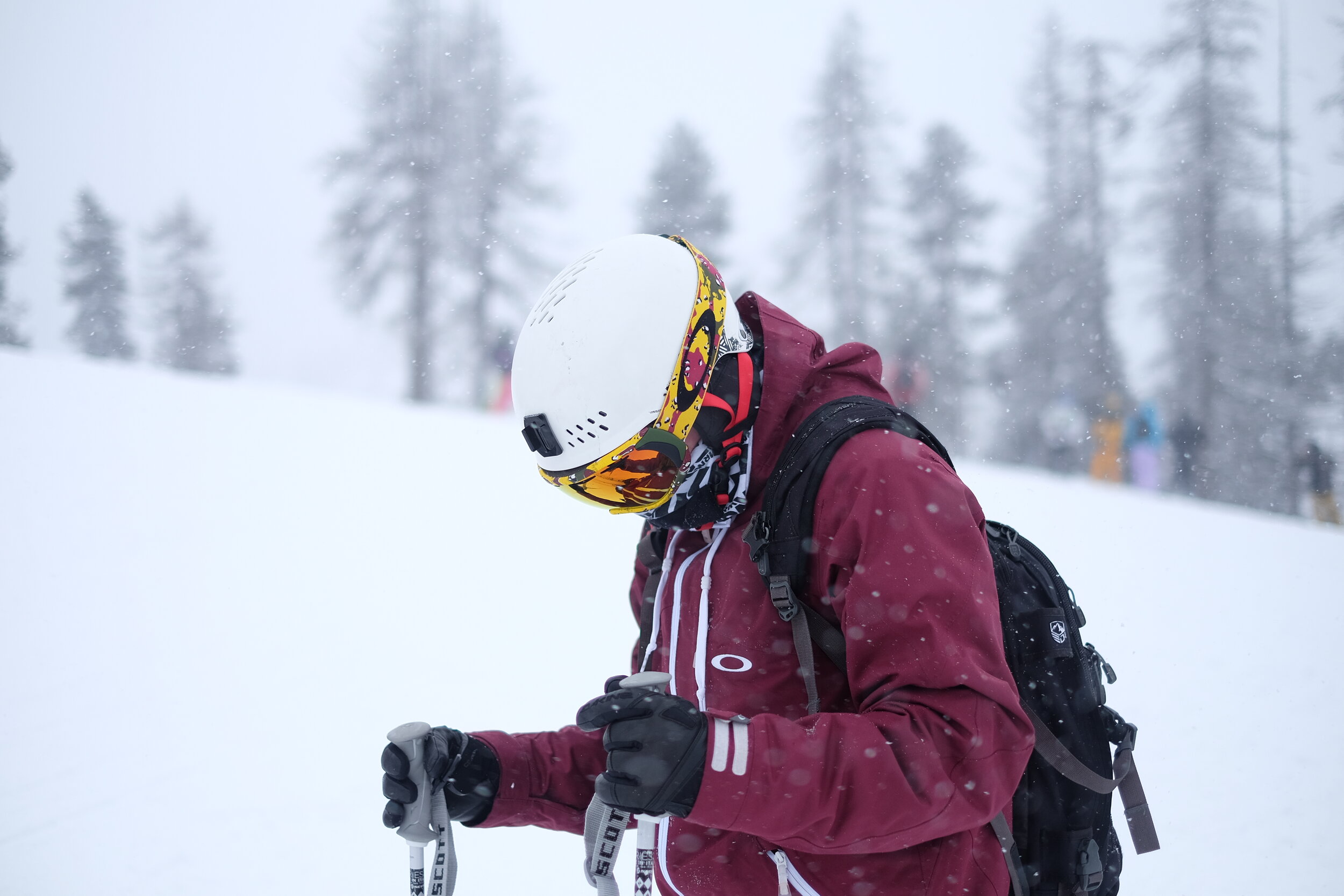 Person in maroon ski jacket and white helmet with colorful goggles holding ski poles in snowy landscape with trees.